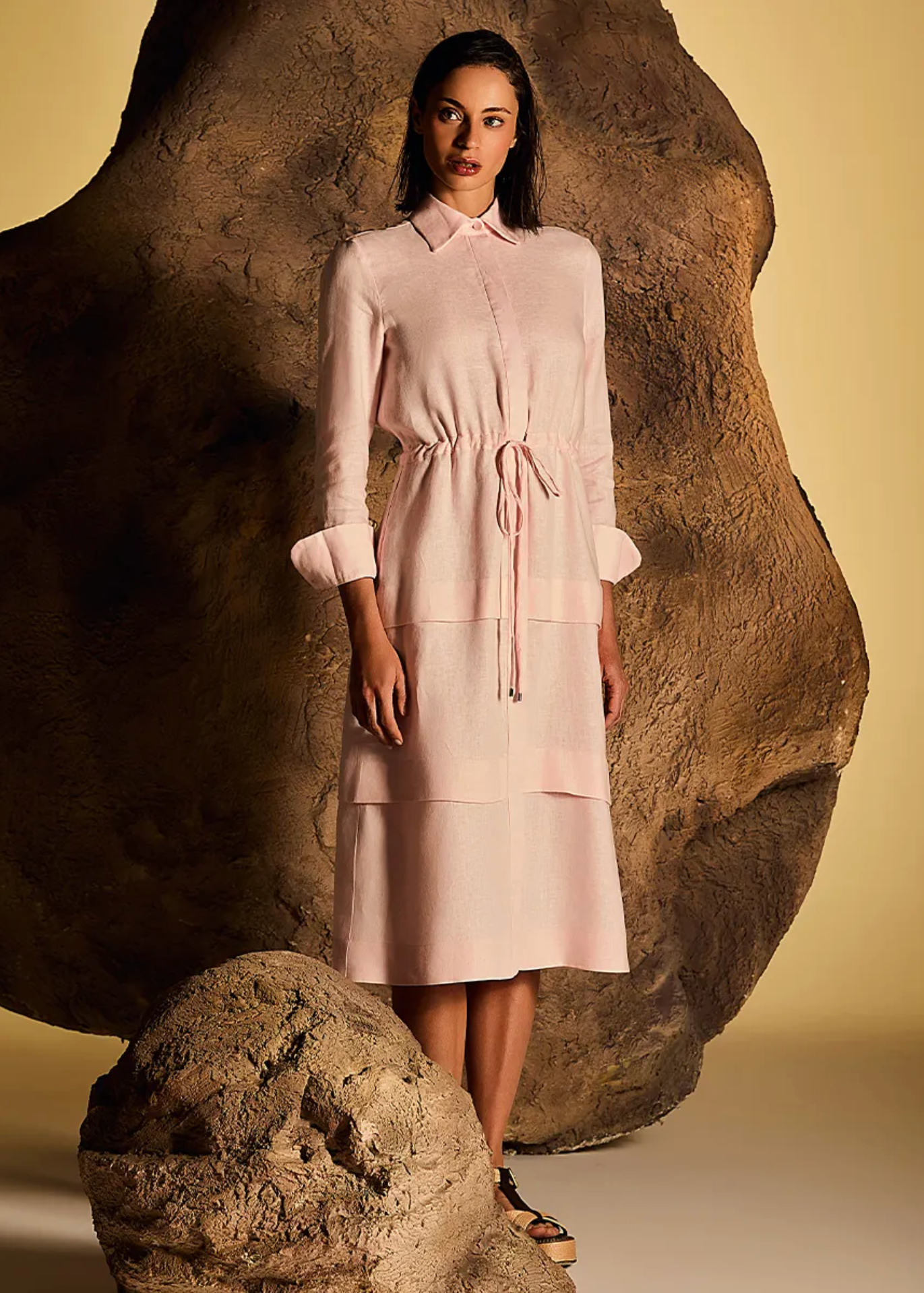 A model stands in front of a large sculptural rock formation wearing a light pink midi shirt dress with three-quarter sleeves, a high collar, and a drawstring waist. The background is a solid warm beige studio backdrop.