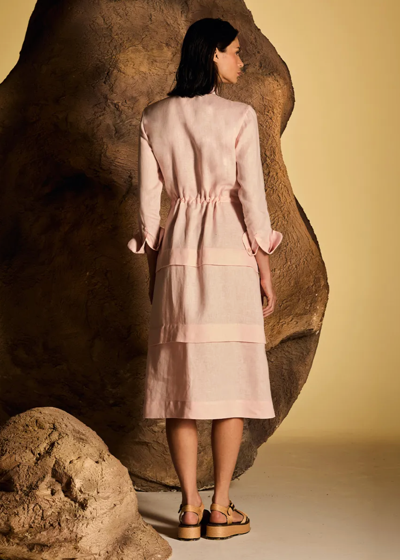 A woman stands with her back to the camera wearing a light pink midi dress with a gathered waist and layered skirt, posed in front of a large textured rock against a warm beige background.