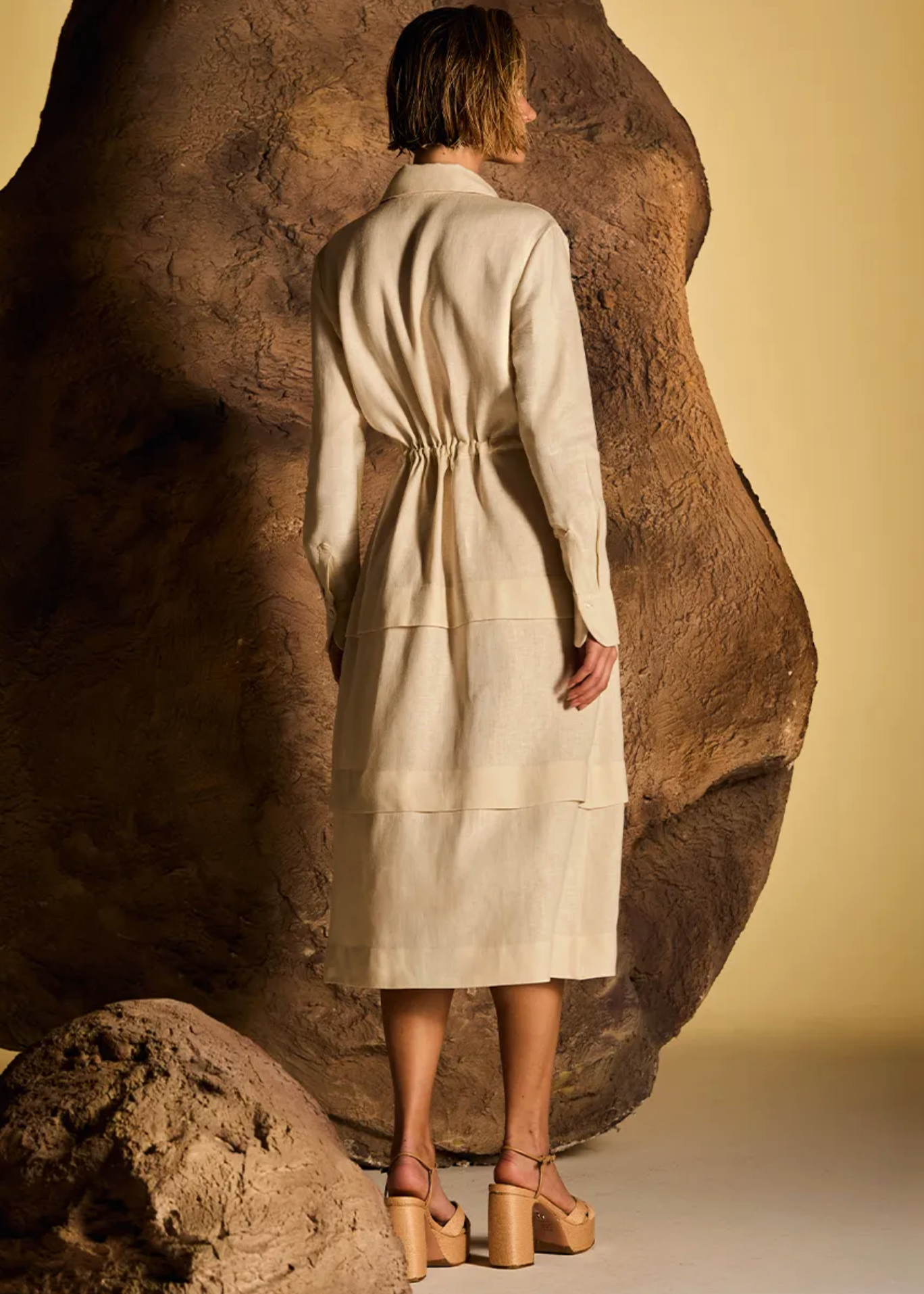 A model stands with their back to the camera wearing a cream-colored linen-blend midi coat with a tie waist, positioned in front of a large sculptural rock against a warm beige background.