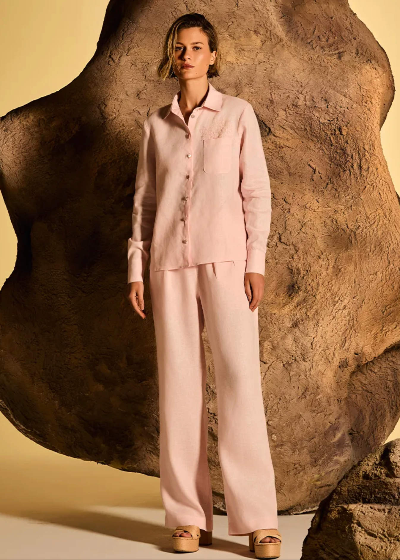A woman stands in front of a large textured rock prop wearing a light blush pink two-piece pajama set with a long-sleeve button-front shirt and matching wide-leg pants, against a warm beige studio backdrop.