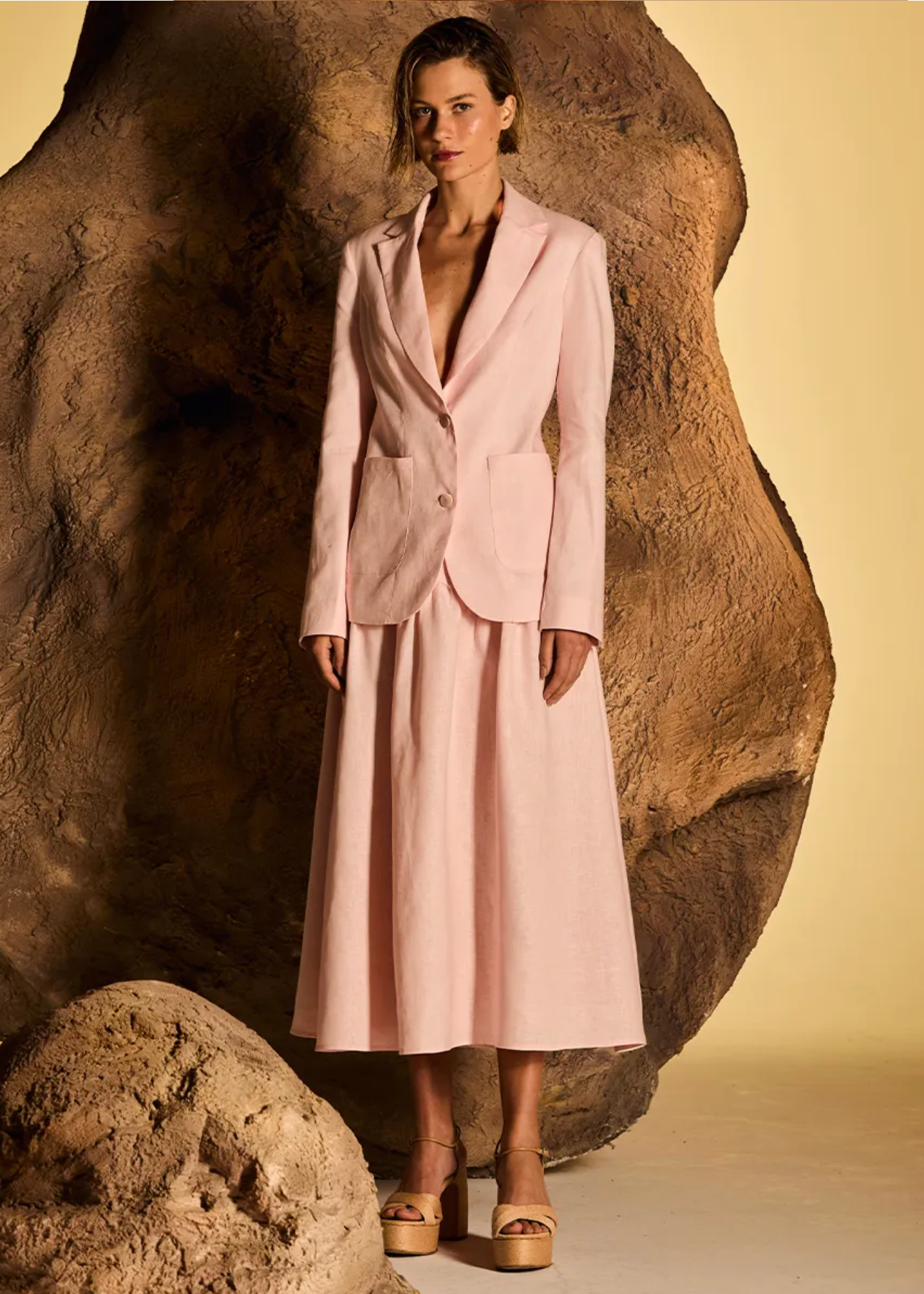 A model stands in front of a large sculptural rock prop wearing a light pink tailored suit set consisting of a blazer and midi skirt, against a warm beige studio backdrop.