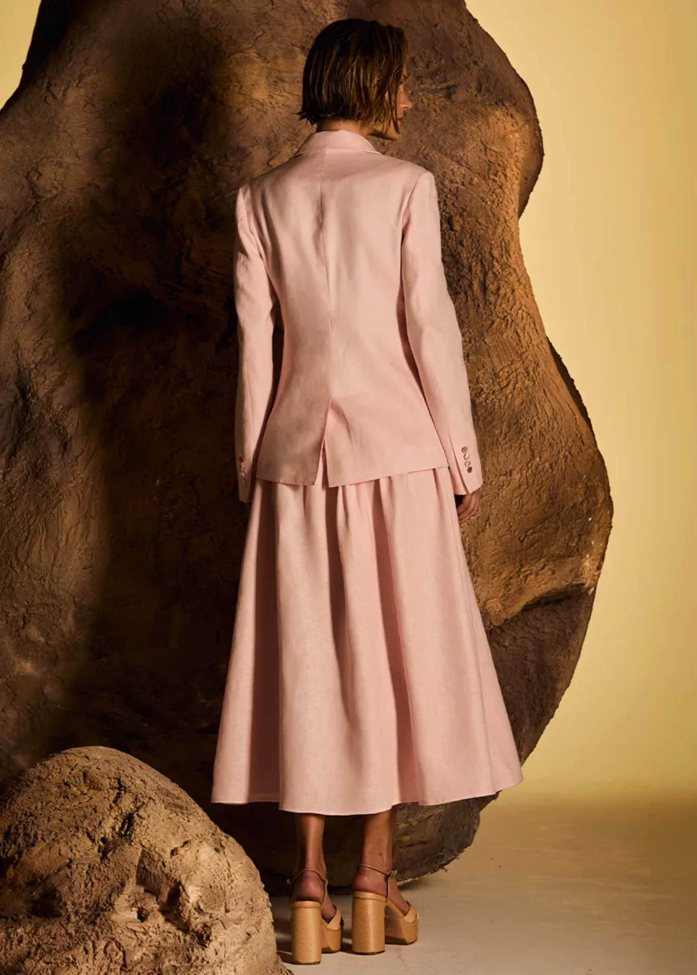 A model stands with her back to the camera wearing a light pink two-piece suit consisting of a tailored blazer and a matching midi skirt, posed in front of a large textured brown rock against a warm beige background.