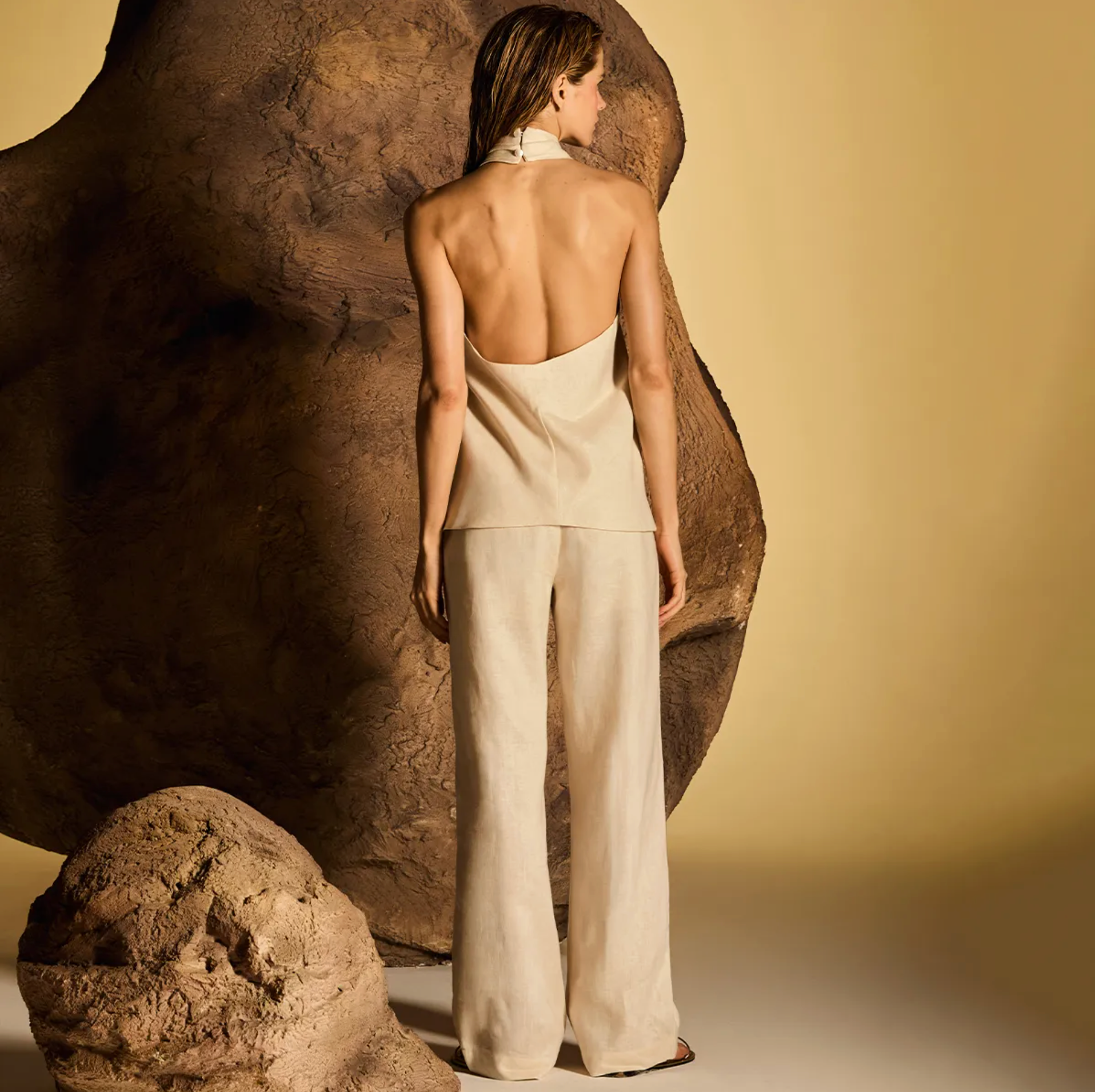 A model stands with her back to the camera wearing a light beige halter top and matching wide-leg trousers, posed in front of a large textured brown rock against a warm beige studio backdrop.