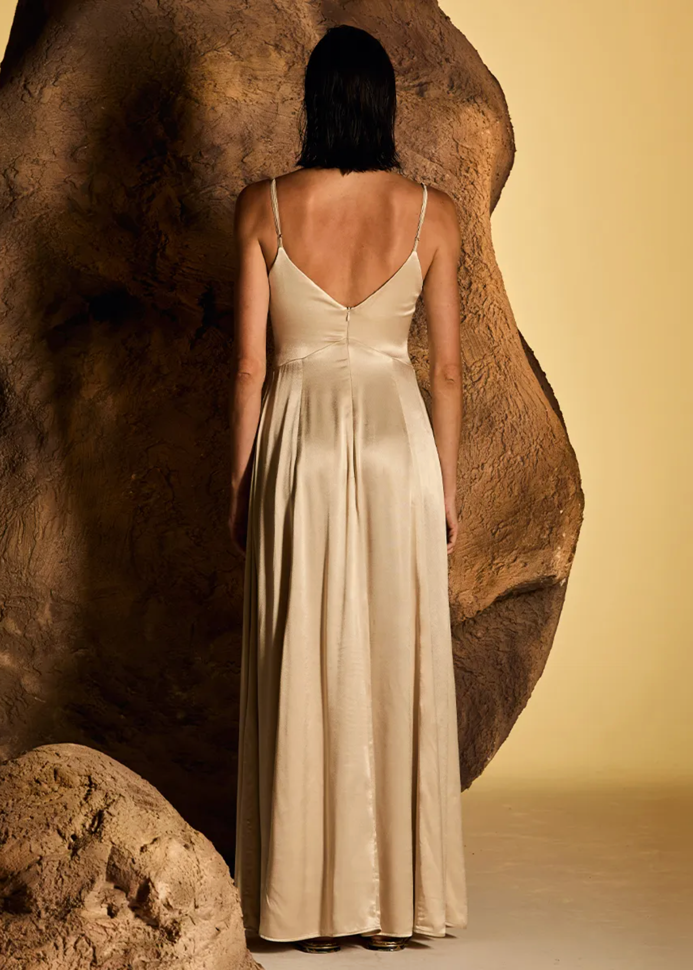 A woman stands with her back to the camera wearing a floor-length satin gown in a warm champagne beige color, posed in front of a large textured rock against a solid beige backdrop.