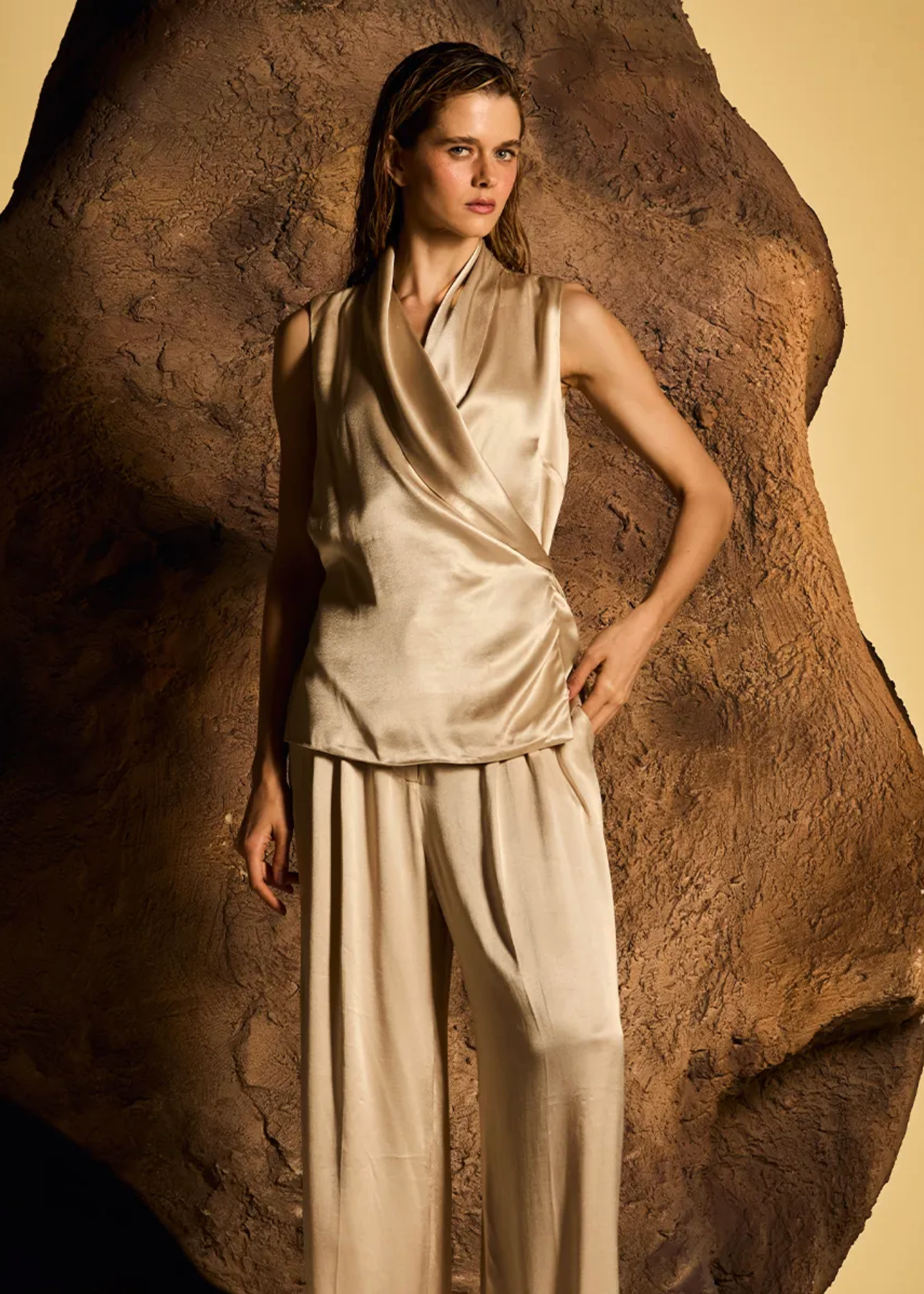 A model wearing a beige satin wrap top and matching wide-leg trousers, standing in front of a large textured brown rock prop against a warm beige background.