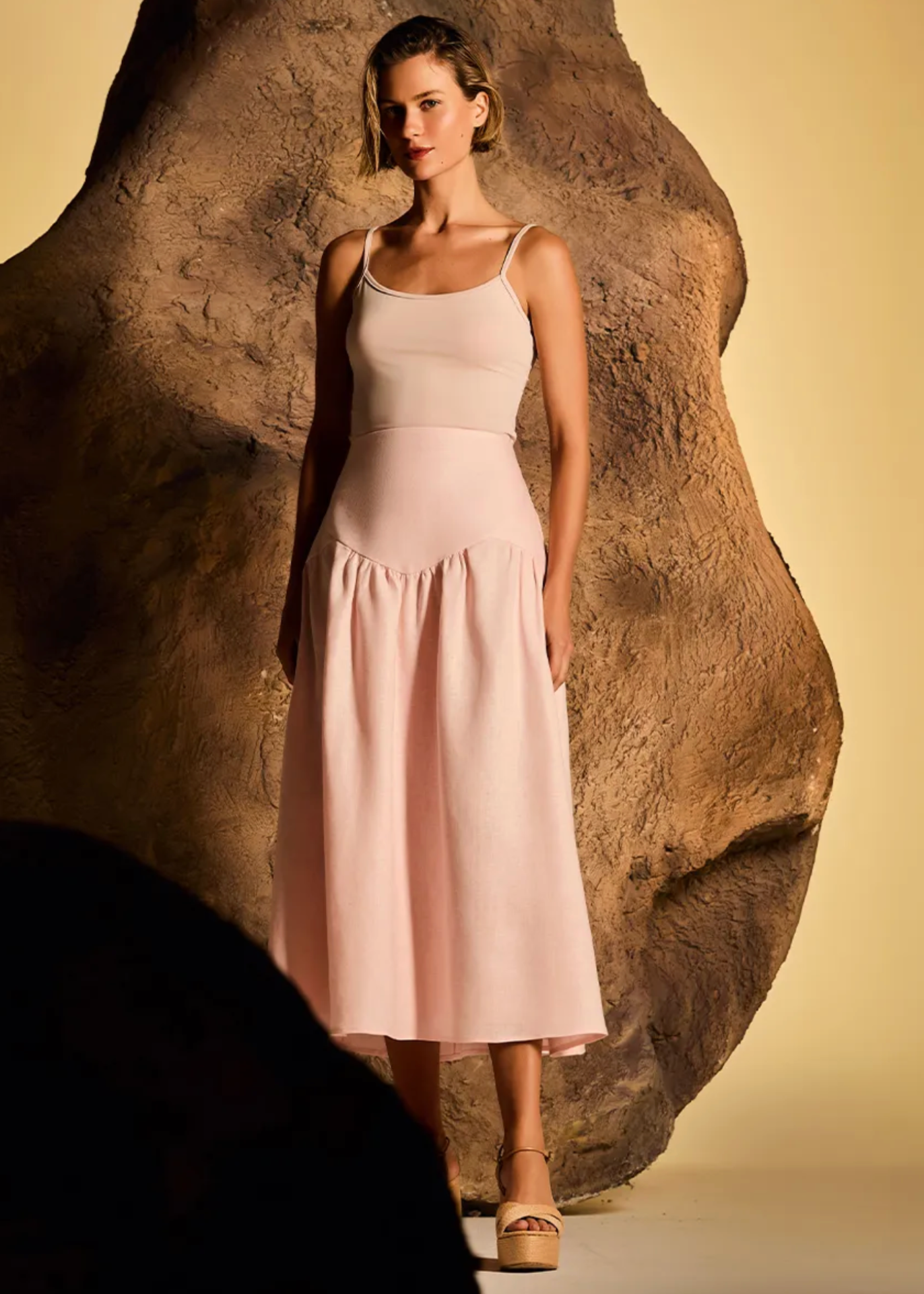 A model wears a light pink sleeveless midi dress with a structured waist and flared skirt, standing in front of a large textured rock prop against a warm beige studio background.
