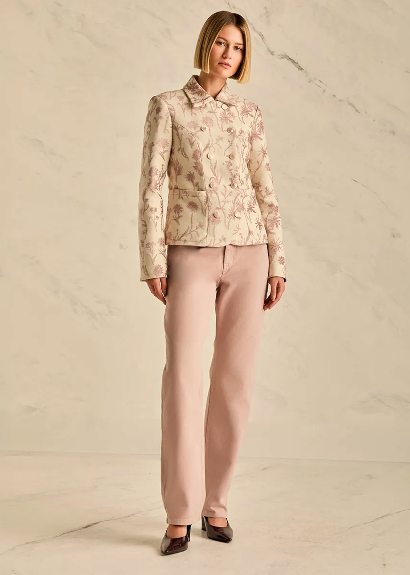 A woman stands against a light marble-textured wall wearing a tailored cream jacket with a muted floral print, light blush pink trousers, and dark pointed flats.