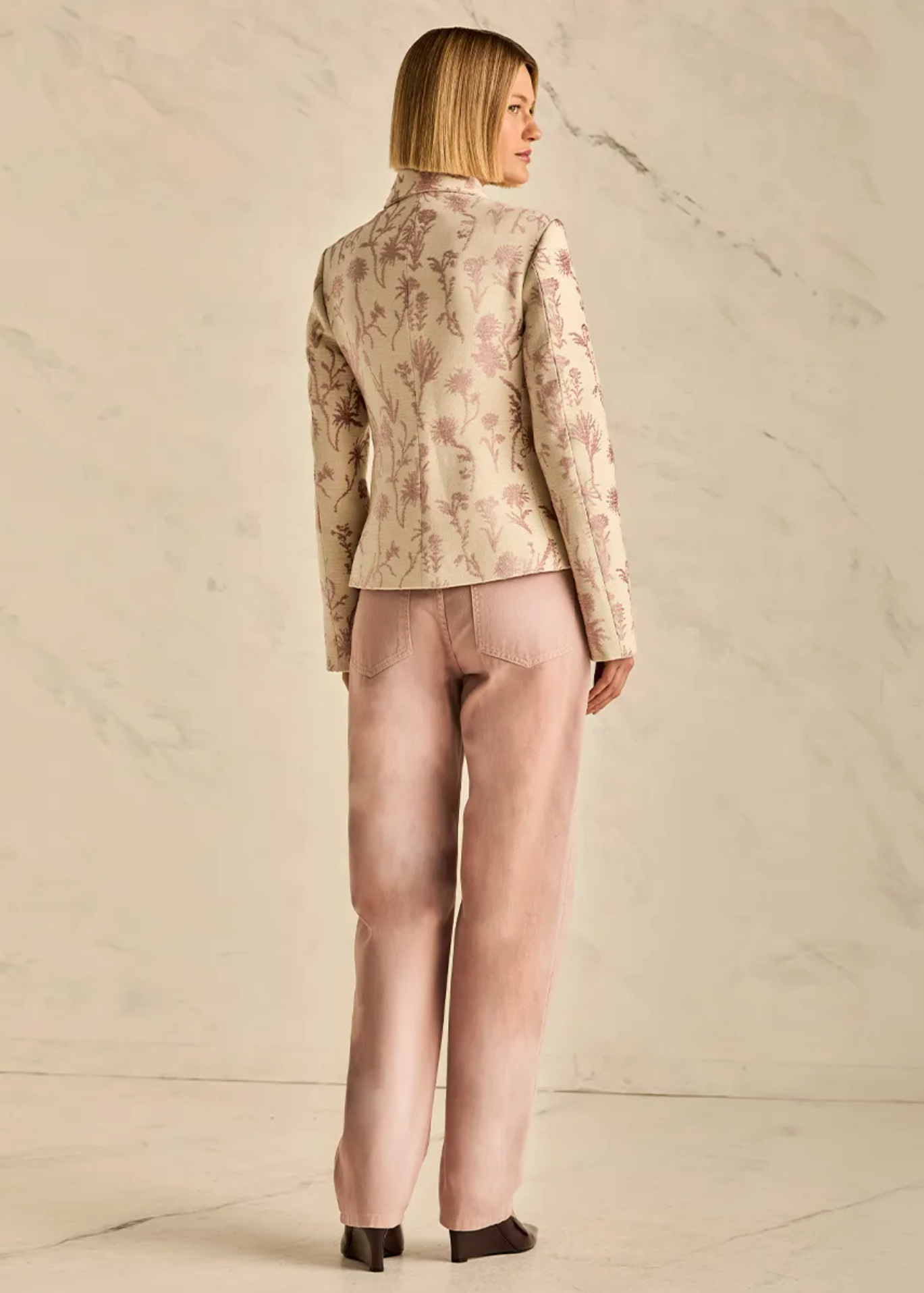 A woman stands with her back to the camera wearing a beige floral-patterned jacket and dusty pink trousers against a light marble wall.