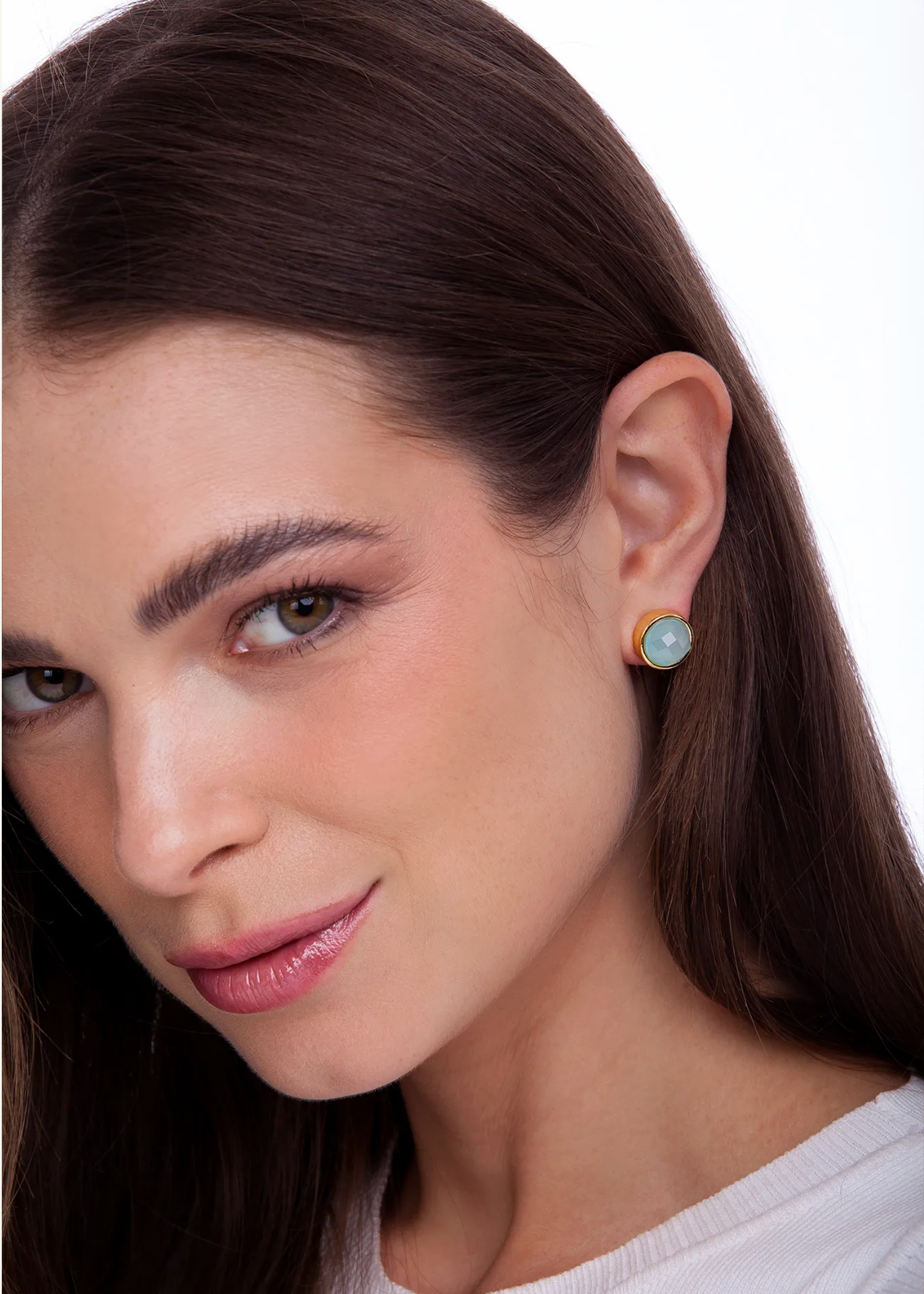 A close-up of a woman wearing a small round stud earring with a light blue-green stone and a gold-toned bezel setting, against a clean white background.