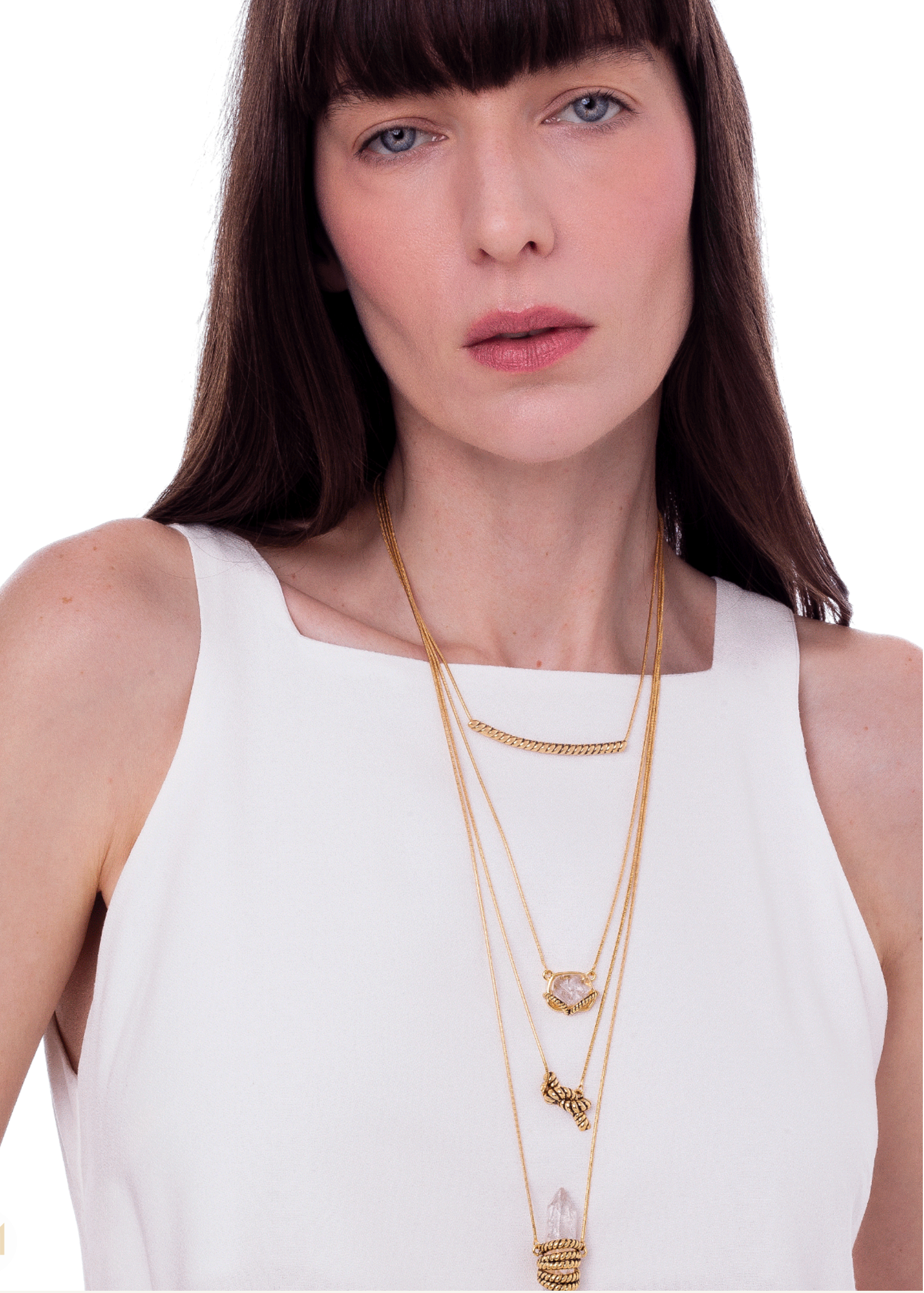 A woman wearing a white sleeveless top and layered gold necklaces, including a delicate chain necklace, a heart-shaped pendant necklace, and a longer necklace with a small cylindrical pendant, against a plain white background.
