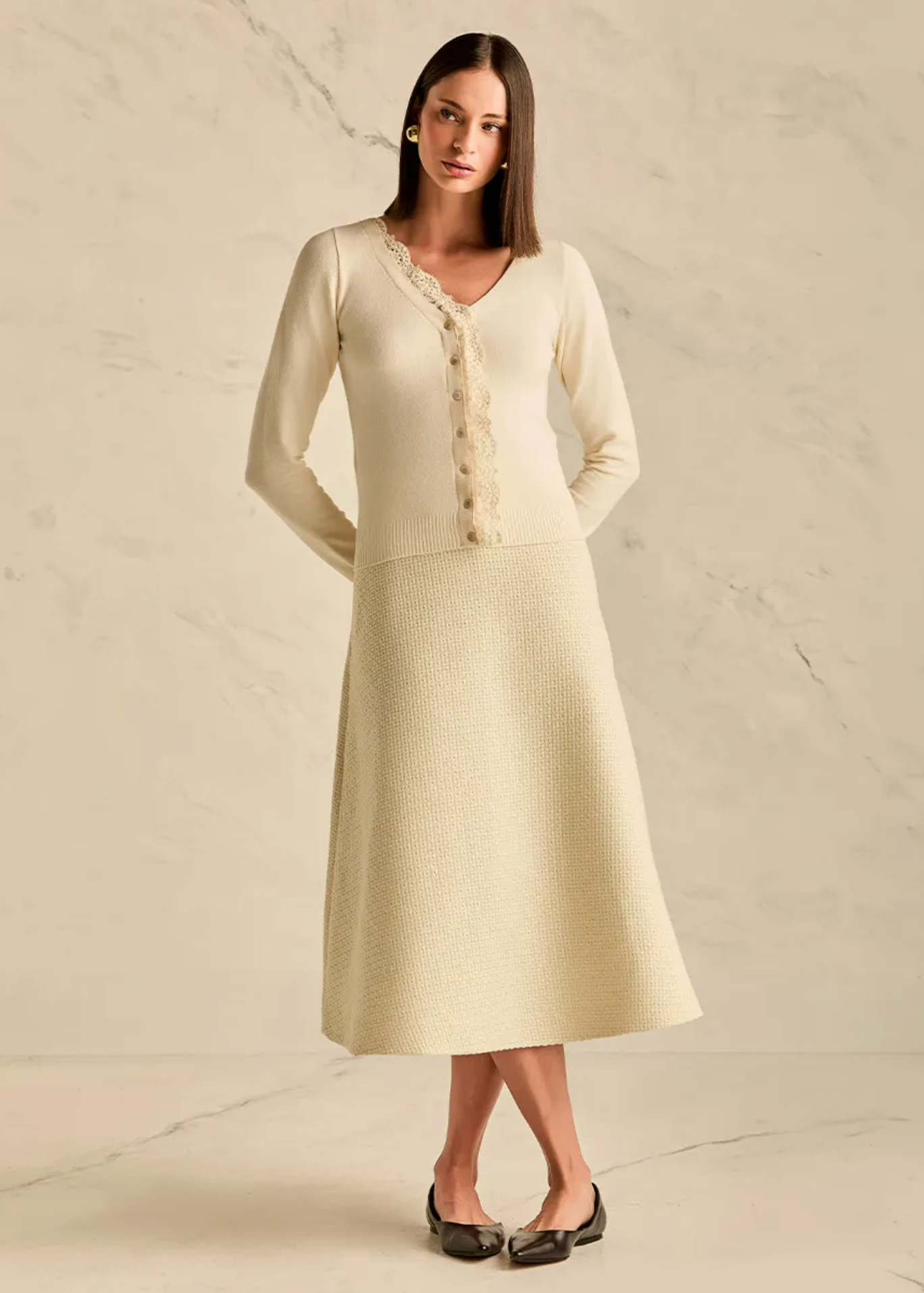 A woman modeling a cream-colored knit midi dress with a textured skirt and a lace-trimmed button-front bodice, standing against a light beige marble wall and floor.