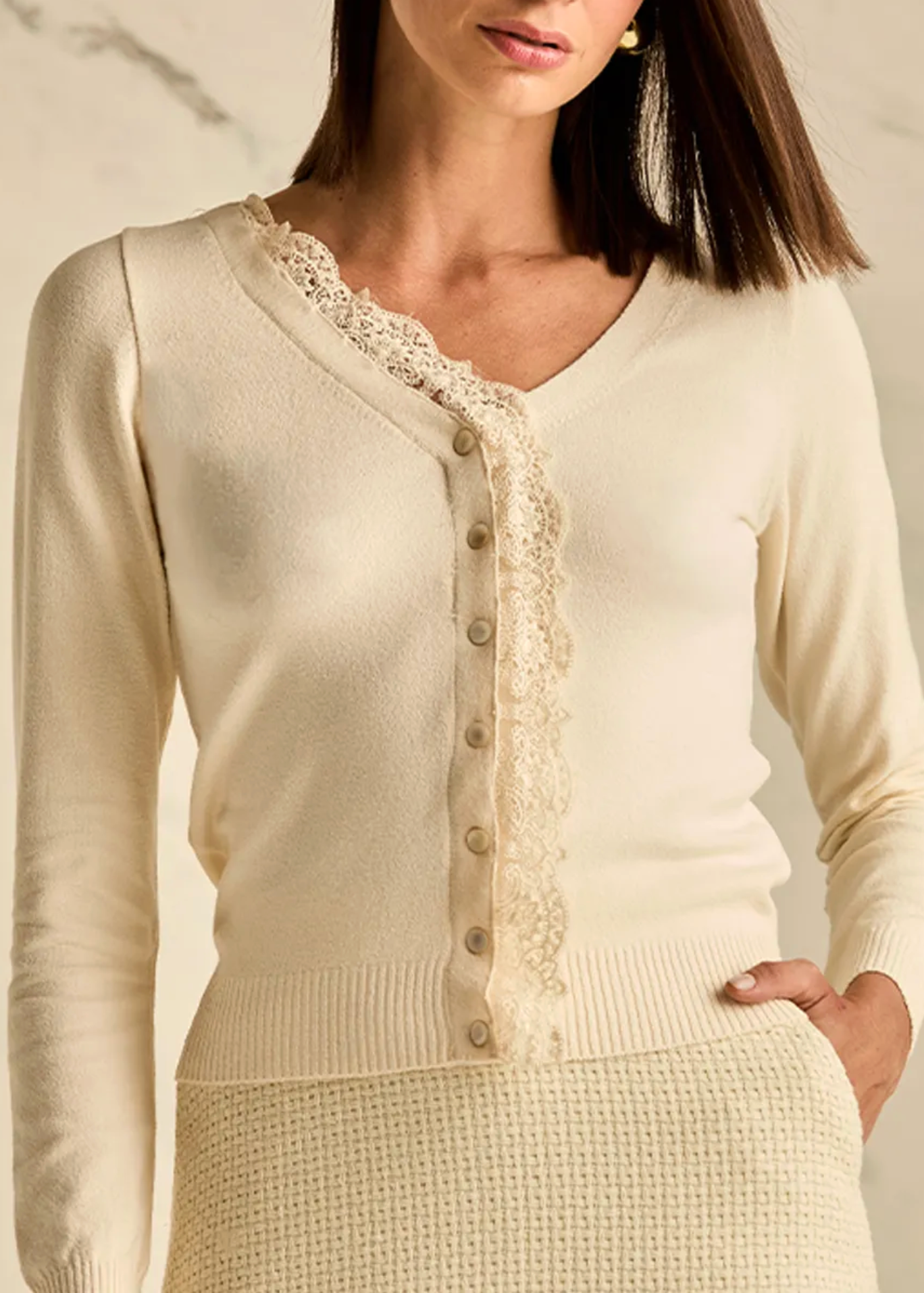 A woman wearing a cream-colored knit cardigan with a lace-trimmed button placket and a matching textured knit skirt, photographed against a light marble wall.
