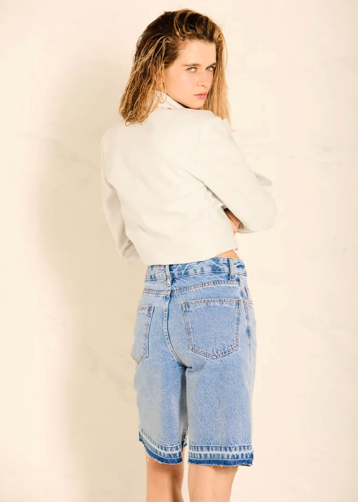 A woman wearing light-wash blue denim shorts and a white cropped sweater, photographed from behind against a plain off-white wall.
