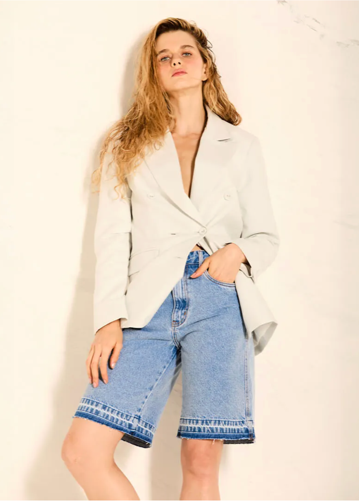 A woman posing in a light gray double-breasted blazer and light-wash denim shorts against a textured off-white wall.