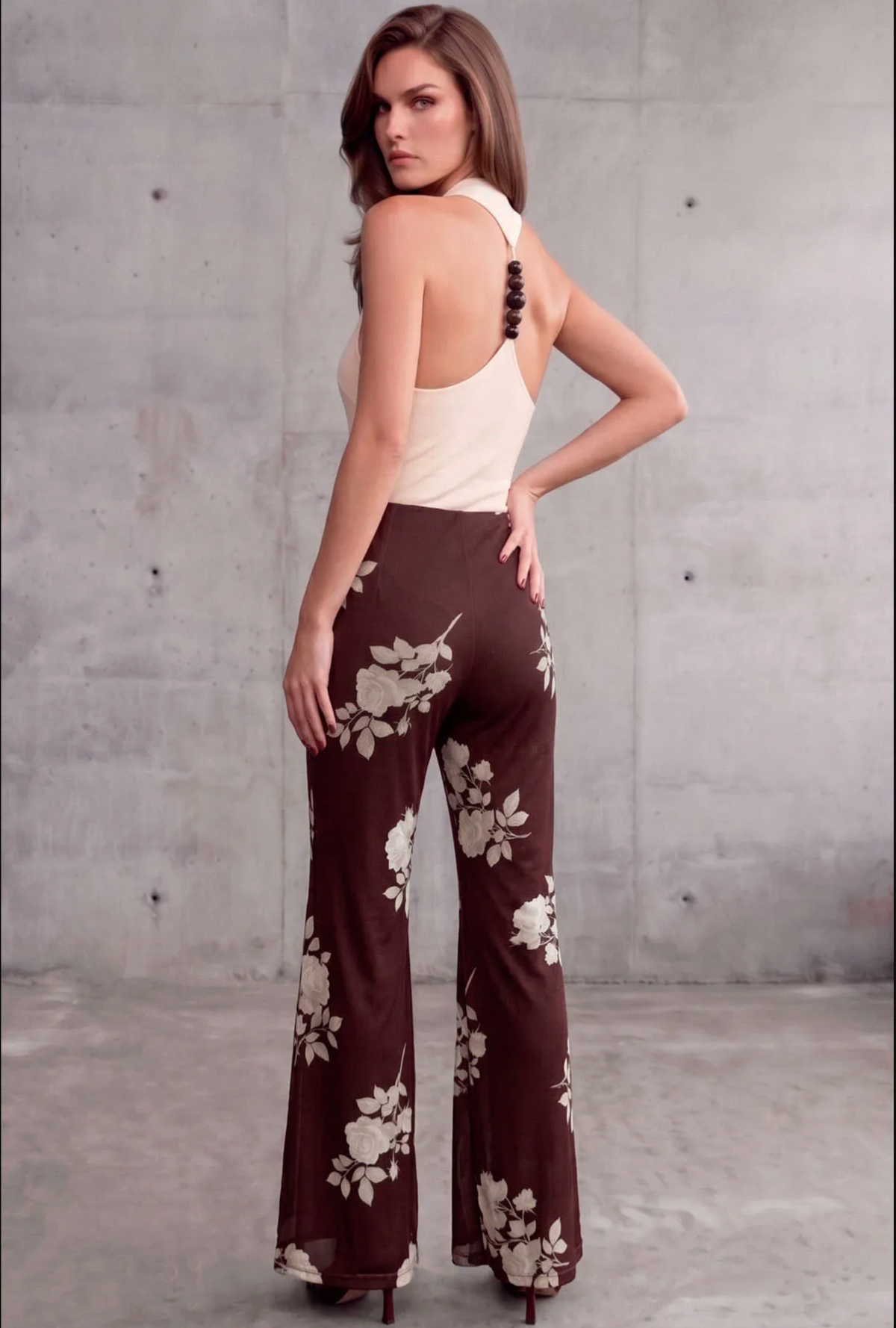 A woman stands with her back to the camera wearing a sleeveless halter-style top in cream and matching brown wide-leg pants with a cream floral print, posed against a concrete wall.