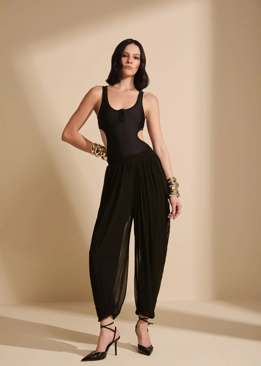 A model wearing a black sleeveless bodysuit with side cutouts and voluminous draped legs, styled with gold bracelets and black heels, set against a warm beige studio backdrop with dramatic shadows.
