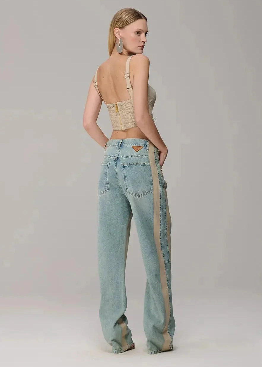 Woman wearing light blue jeans with a beige stripe and a beige top on a plain background