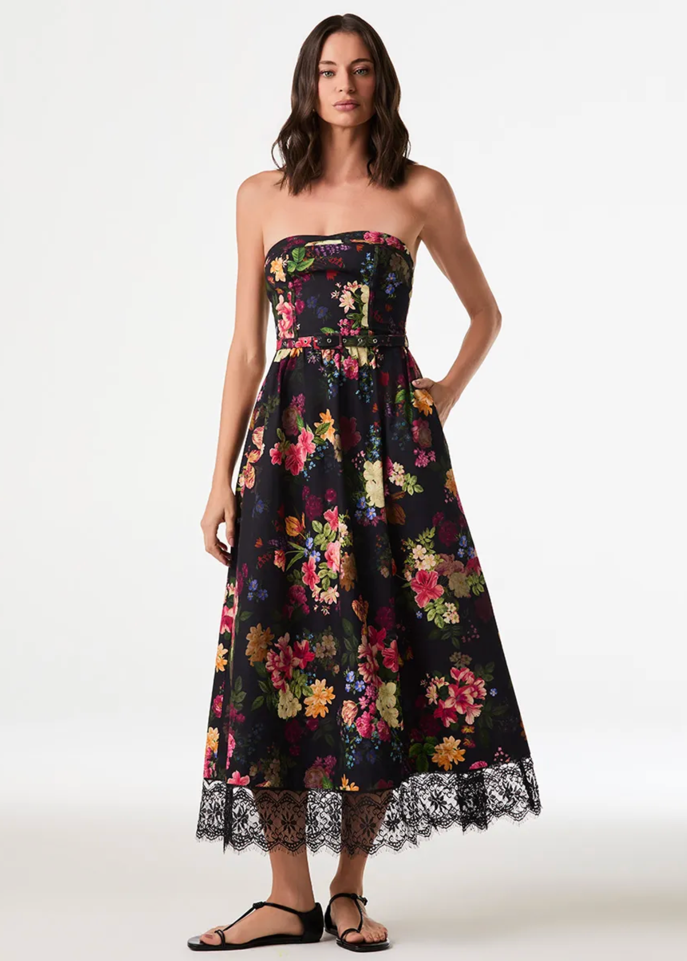 A woman modeling a strapless midi dress with a black background and a vibrant multicolor floral print, featuring a lace-trimmed hem and black sandals, against a plain white studio background.