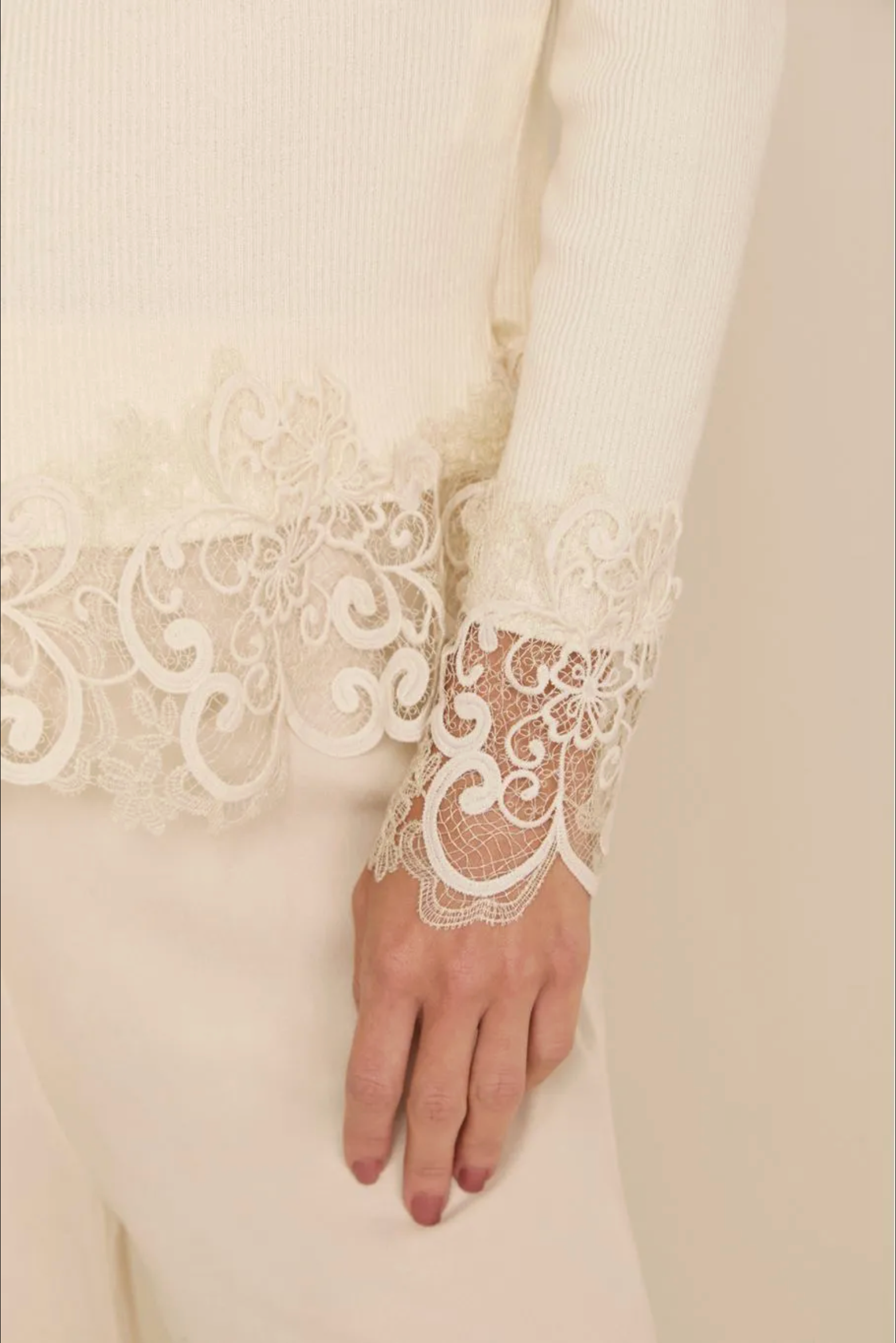 A close-up of a person's hand wearing a cream ribbed knit top with ornate floral lace cuffs, paired with matching cream trousers against a soft beige background.
