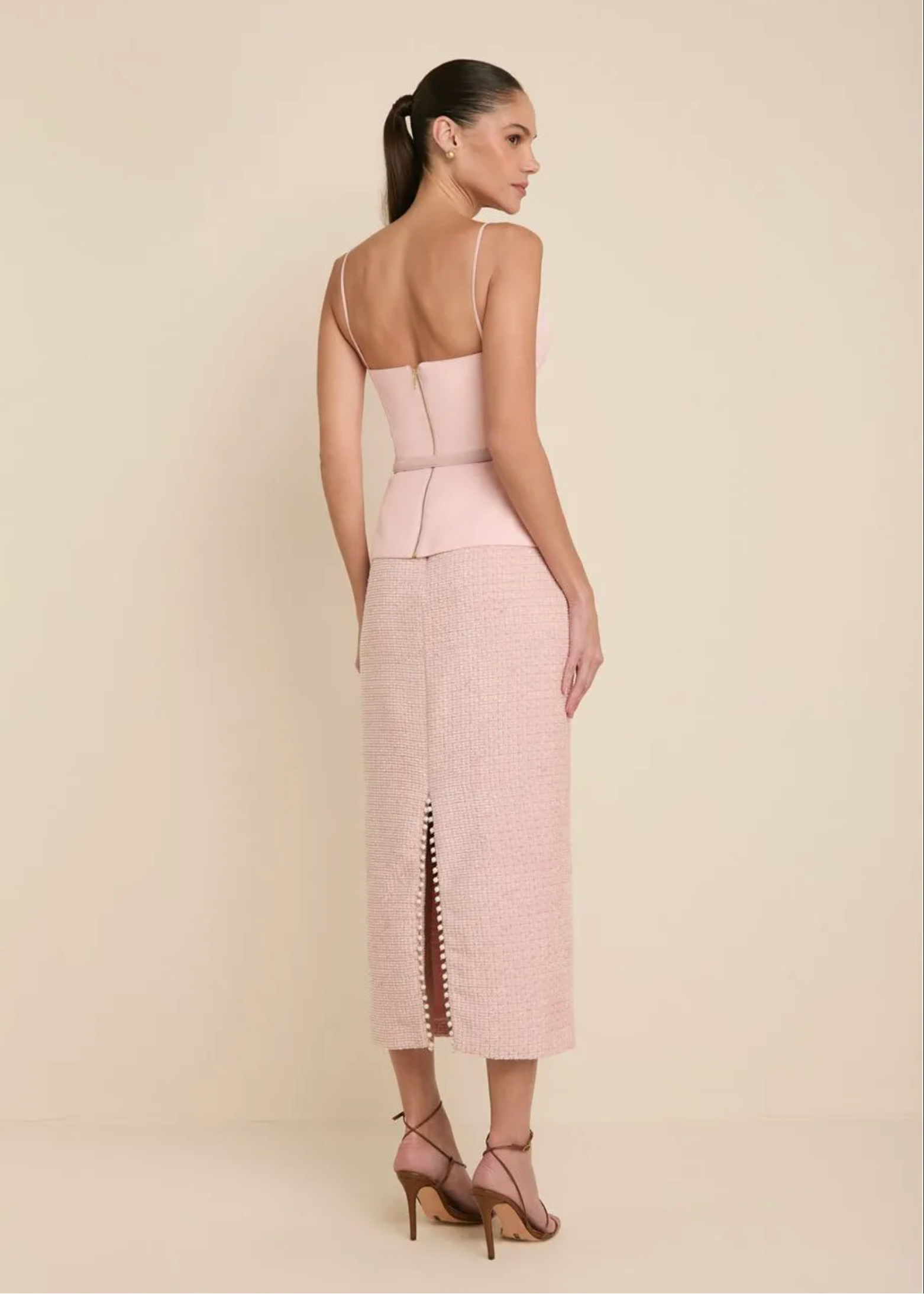 A woman is shown from the back wearing a light pink sleeveless dress with a structured corset-style bodice and a textured midi skirt with a front slit, paired with matching pink strappy heels, against a solid beige studio background.