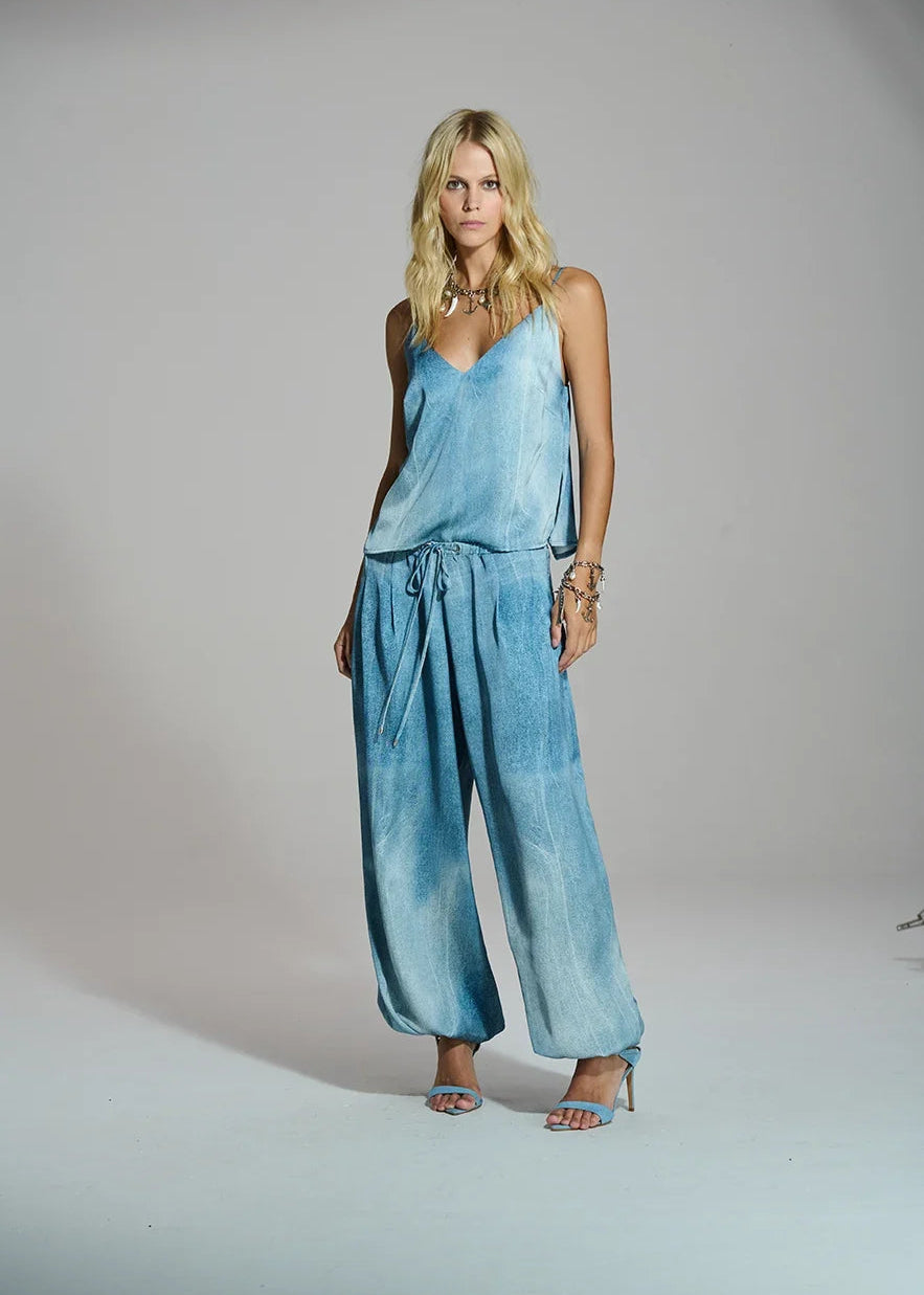 A blonde model wearing a light blue tie-dye sleeveless jumpsuit with wide-leg pants and matching sandals, standing against a neutral gray studio background.