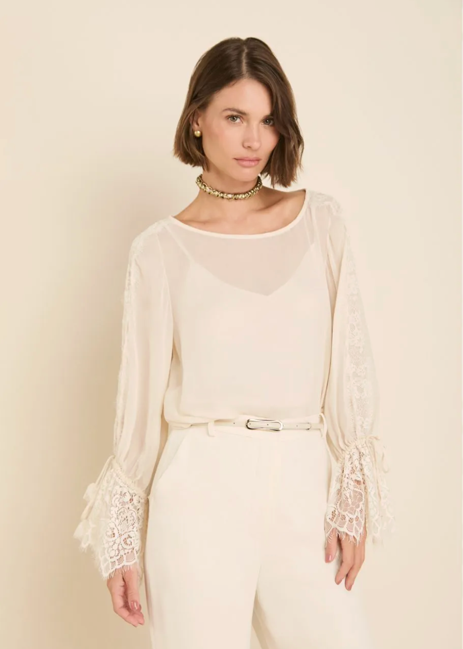 A woman modeling a cream-colored long-sleeve blouse with sheer mesh panels and intricate lace detailing on the sleeves, paired with cream trousers and a gold choker necklace, set against a solid light beige background.