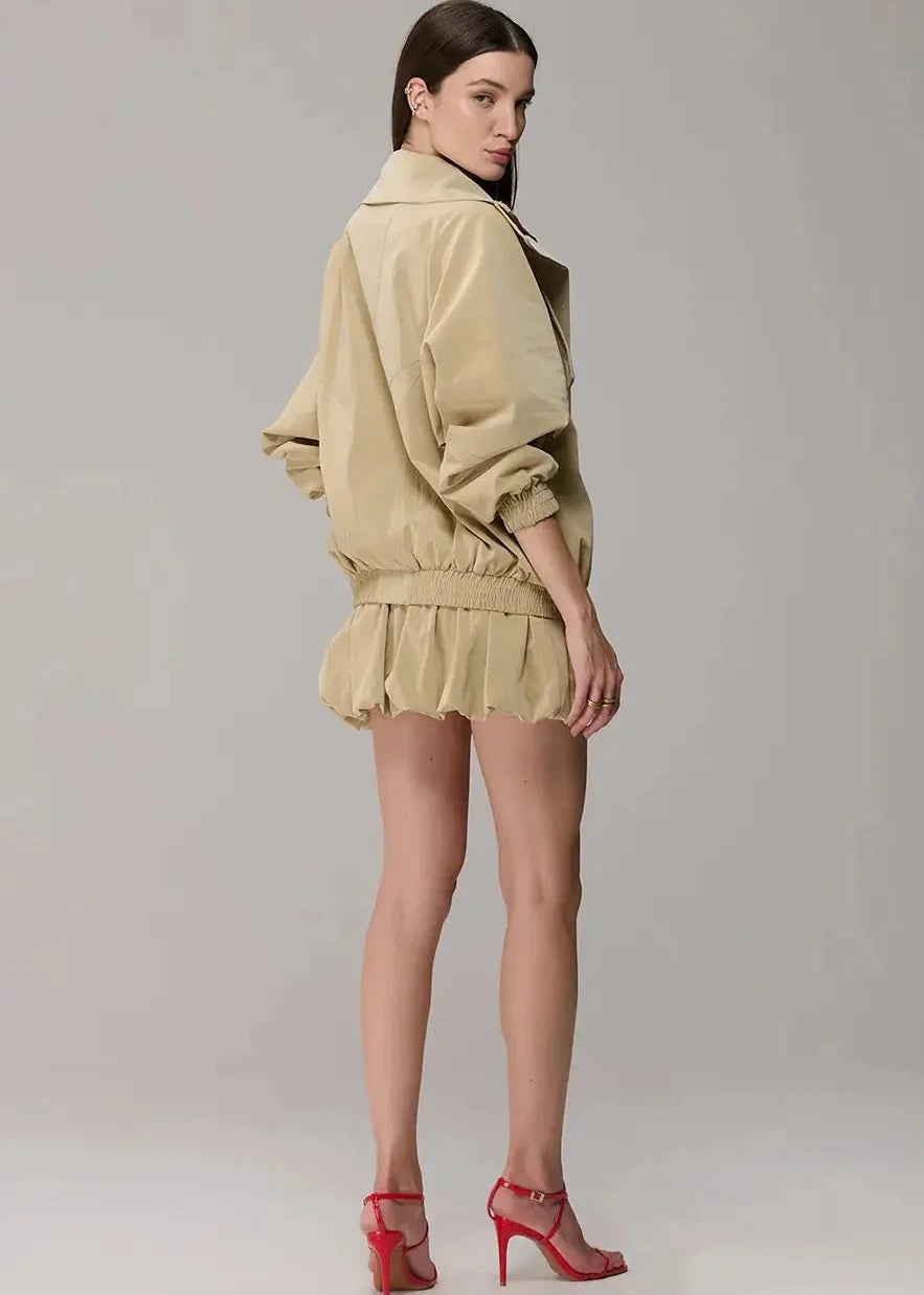A model is shown from the back wearing a beige oversized jacket and matching ruffled mini skirt, paired with red high-heeled sandals, against a neutral gray studio background.