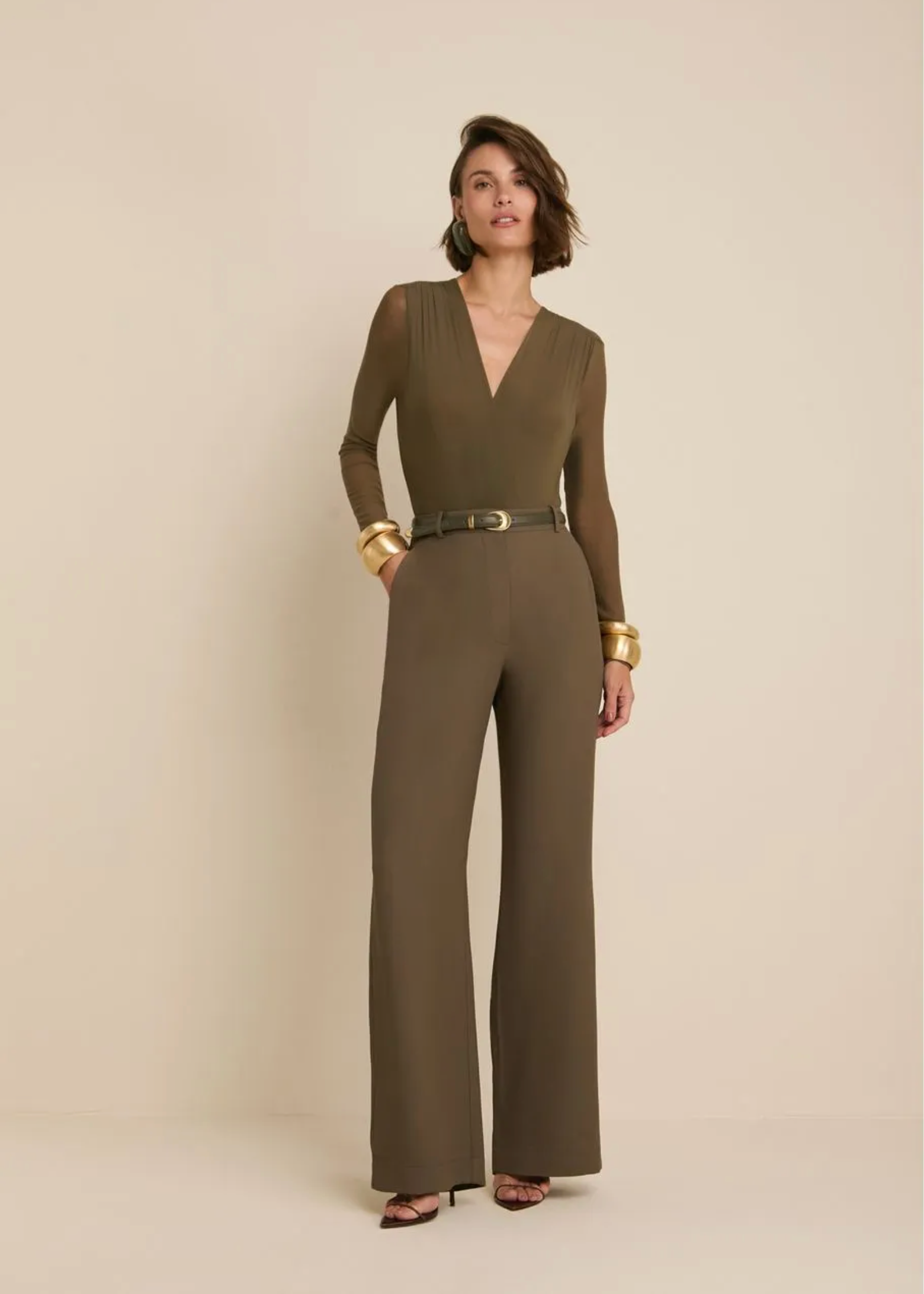 A woman modeling a long-sleeve olive green wide-leg jumpsuit with a matching belt, styled with gold bracelets, against a plain beige studio background.
