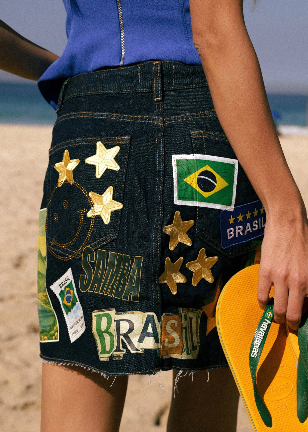 Person wearing a denim skirt with Brazilian-themed patches on a beach.
