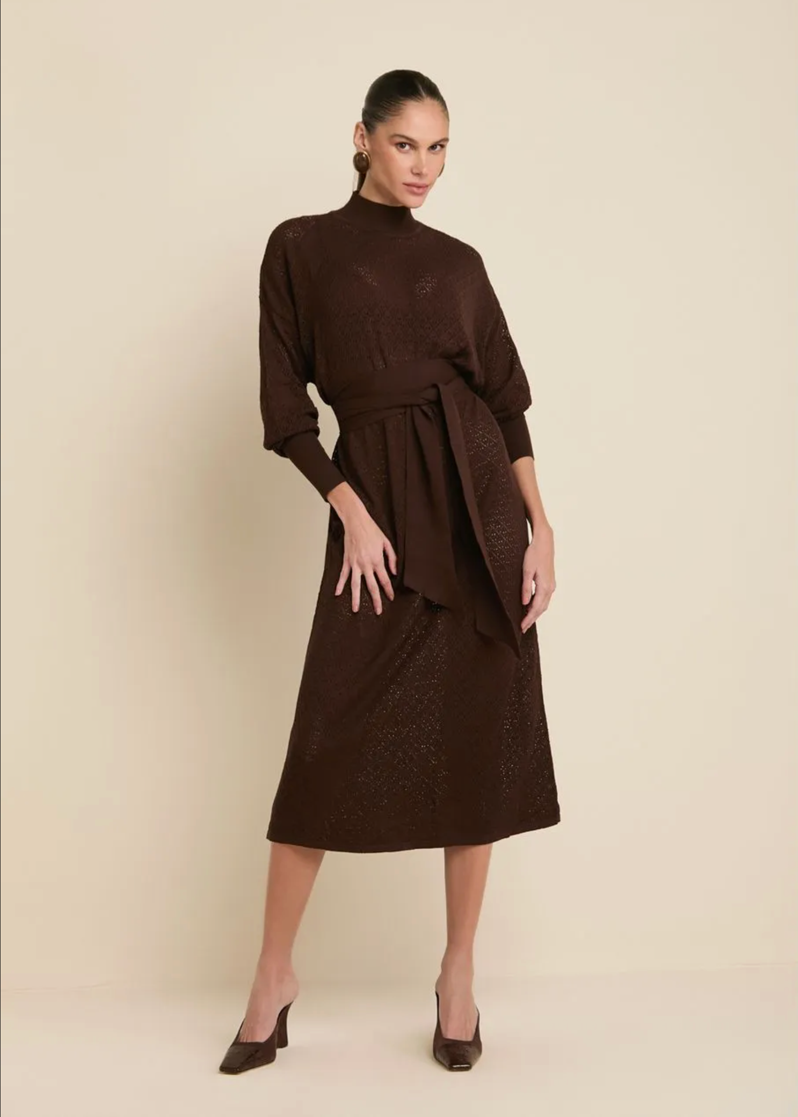 A woman modeling a dark brown midi dress with a mock neck, three-quarter sleeves, and a matching self-tie belt, set against a solid light beige studio background.