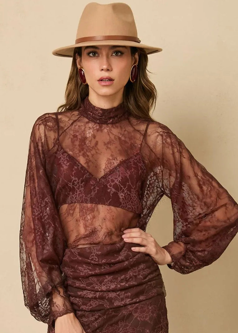 Woman wearing a sheer maroon top and pants with a beige hat on a beige background