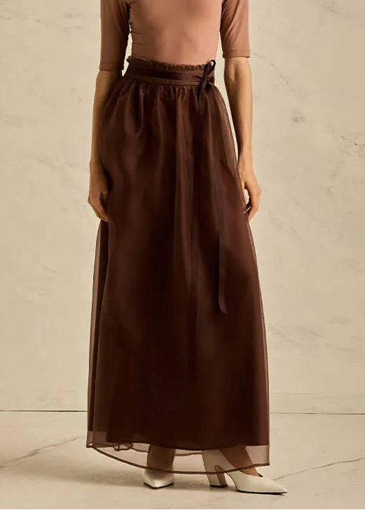 Woman wearing a brown long-sleeve top and long skirt against a neutral background