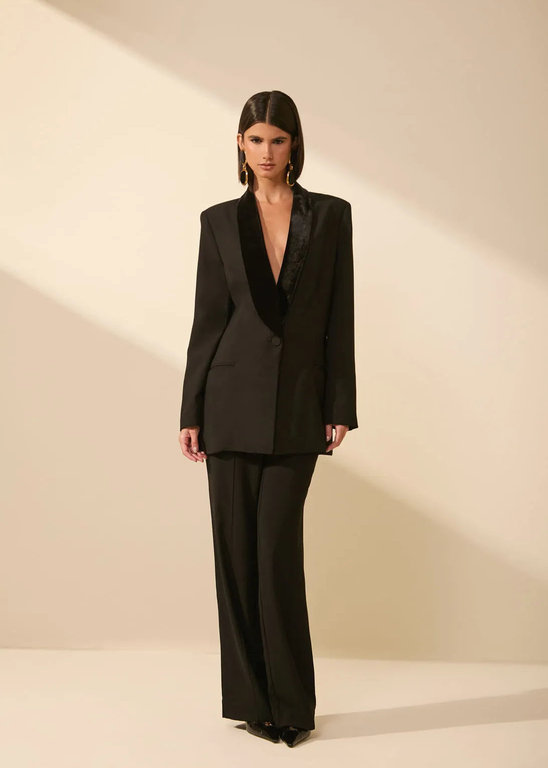 A model stands in a studio wearing a black tailored suit with a structured blazer featuring a wide lapel and matching wide-leg trousers, set against a warm beige background with dramatic diagonal lighting.