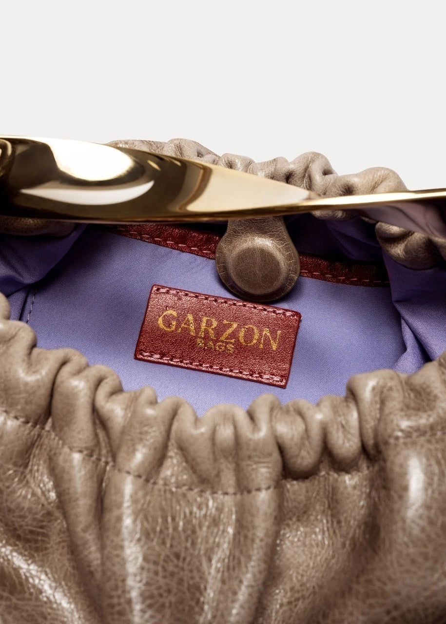 Close-up of a brown leather bag with a gold handle and 'Garzon' brand label on a purple fabric background.
