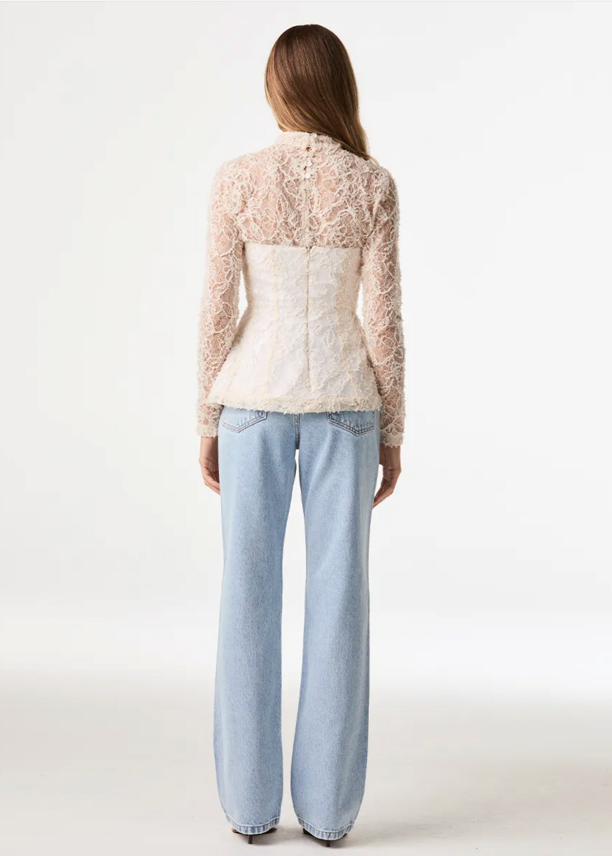 A model viewed from the back wearing a cream lace long-sleeve top with a structured corset-style waist and light-wash wide-leg jeans against a plain white studio background.