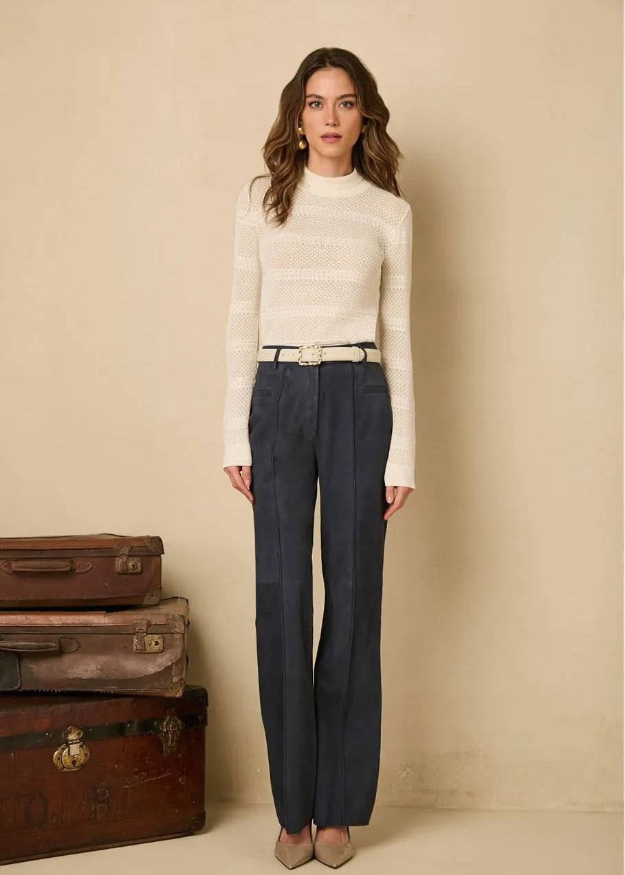 Woman wearing a white sweater and dark pants standing in front of vintage suitcases against a beige wall.