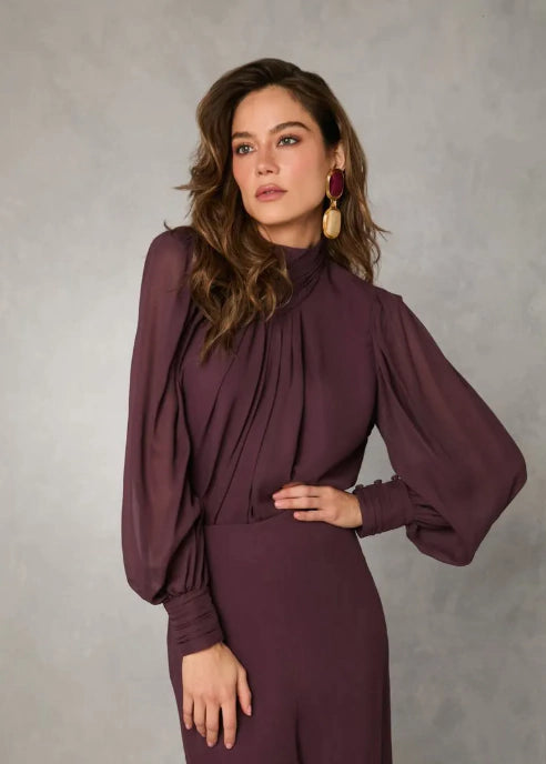 A woman modeling a high-neck, long-sleeved plum purple dress with voluminous balloon sleeves and gathered detailing at the chest, posed against a textured gray studio backdrop.