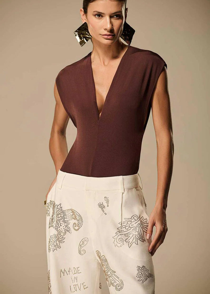 Woman wearing a brown top and white pants with embroidery, standing against a beige background