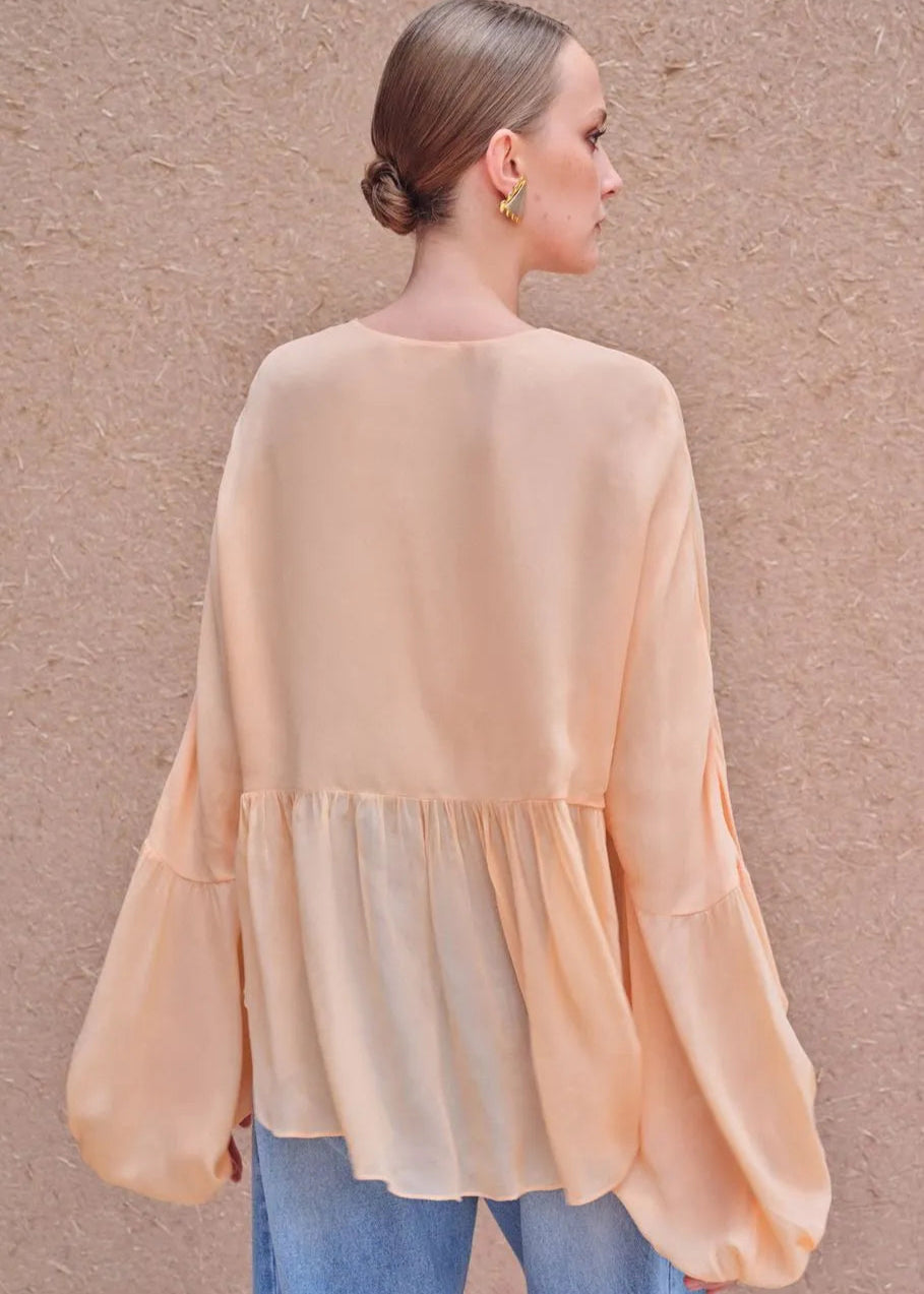 Woman wearing a peach-colored blouse with long sleeves against a beige wall.