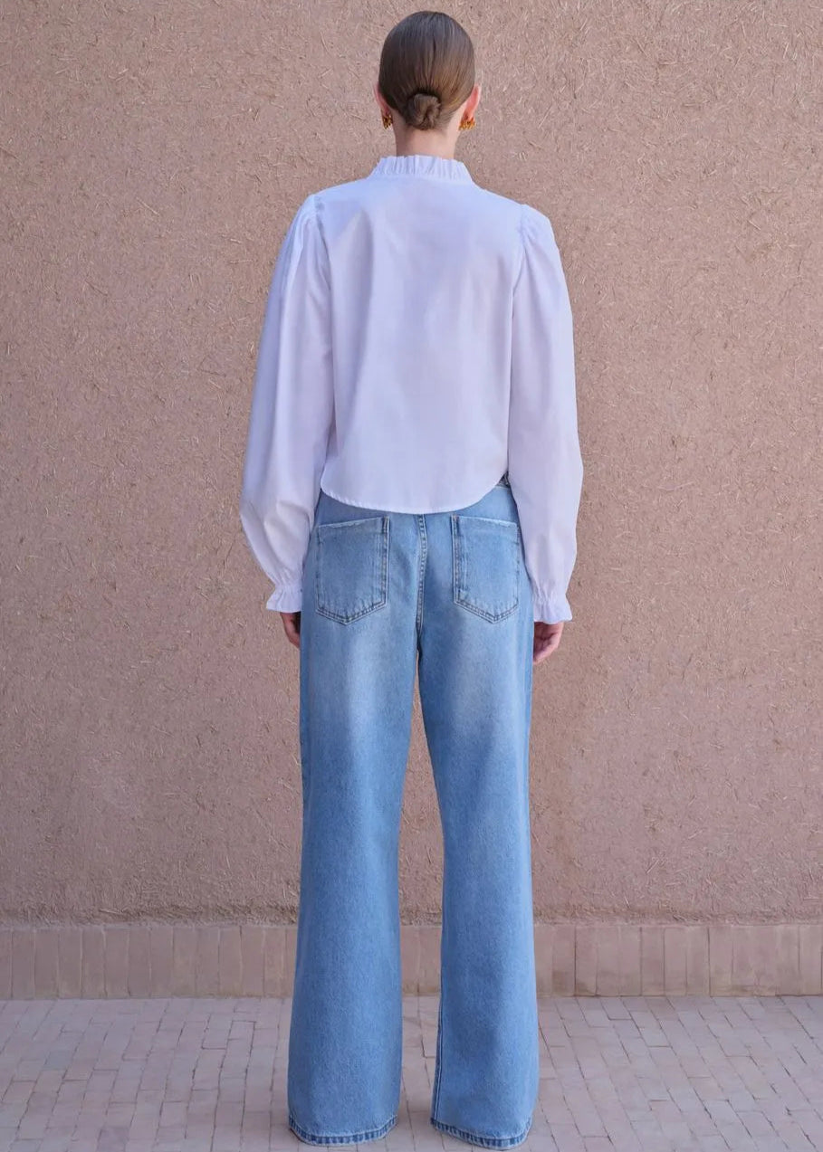 Person wearing a light blue shirt and jeans standing against a beige wall.