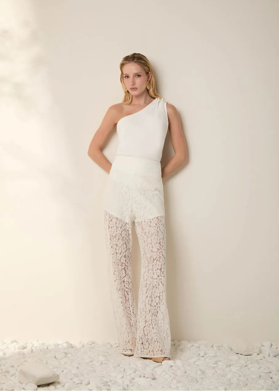 Woman wearing a white one-shoulder top and lace pants against a light background
