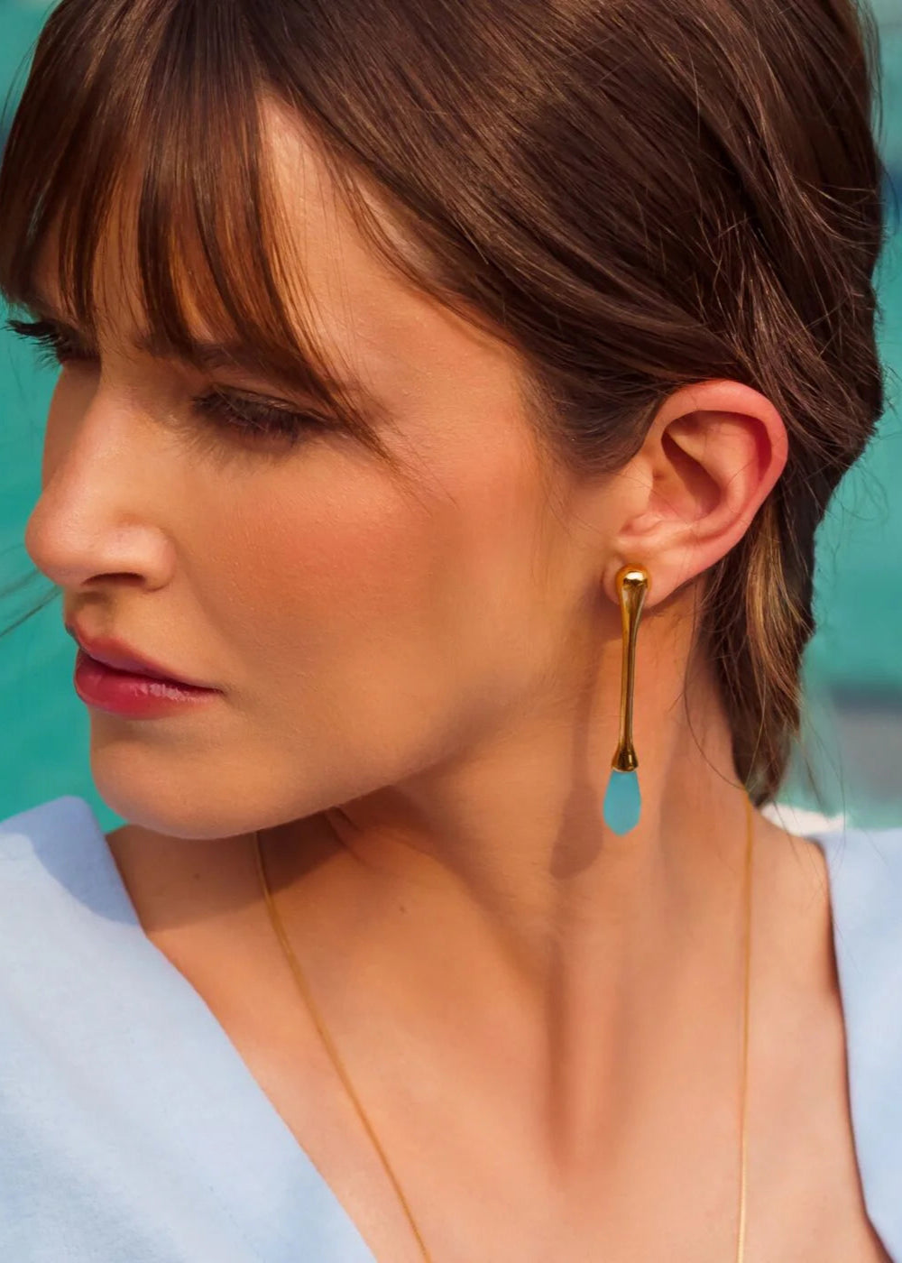 Woman wearing a gold earring with a blue teardrop pendant against a blurred outdoor background.