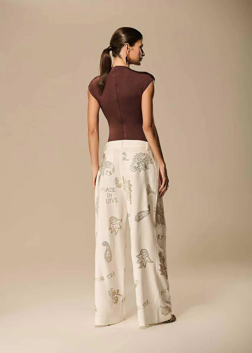Woman wearing a brown top and white pants with embroidery on a beige background