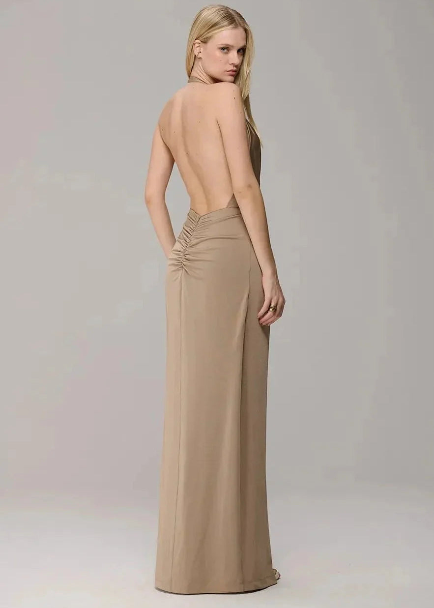 A woman modeling a floor-length beige gown from the back, featuring a deep open back and gathered detailing at the waist, set against a plain light gray studio background.