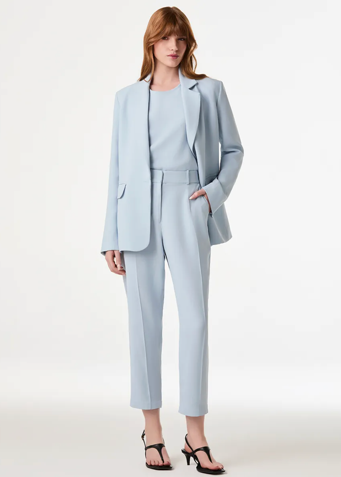 A model wearing a light blue tailored suit set consisting of a blazer, matching top, and cropped trousers, paired with black strappy heels against a plain light background.
