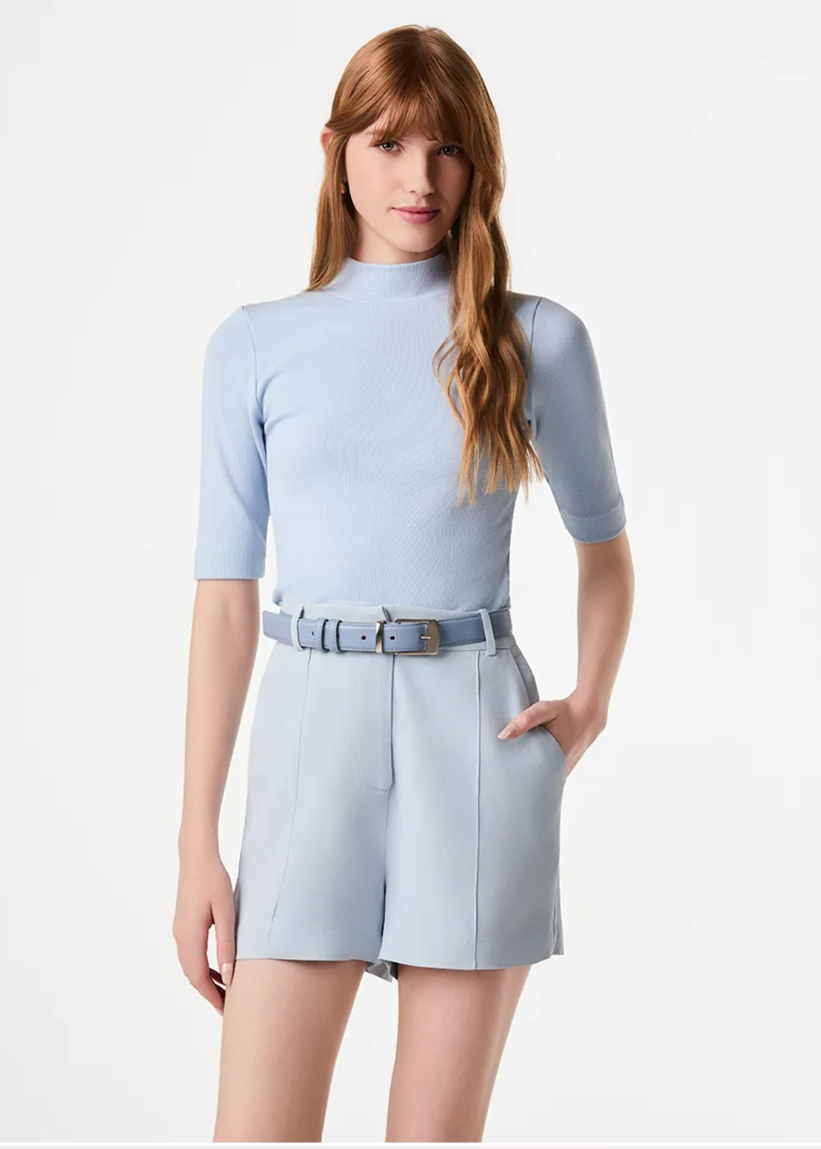 A woman modeling a light blue ribbed short-sleeve mock-neck top paired with matching light blue shorts and a matching belt, set against a plain white studio background.