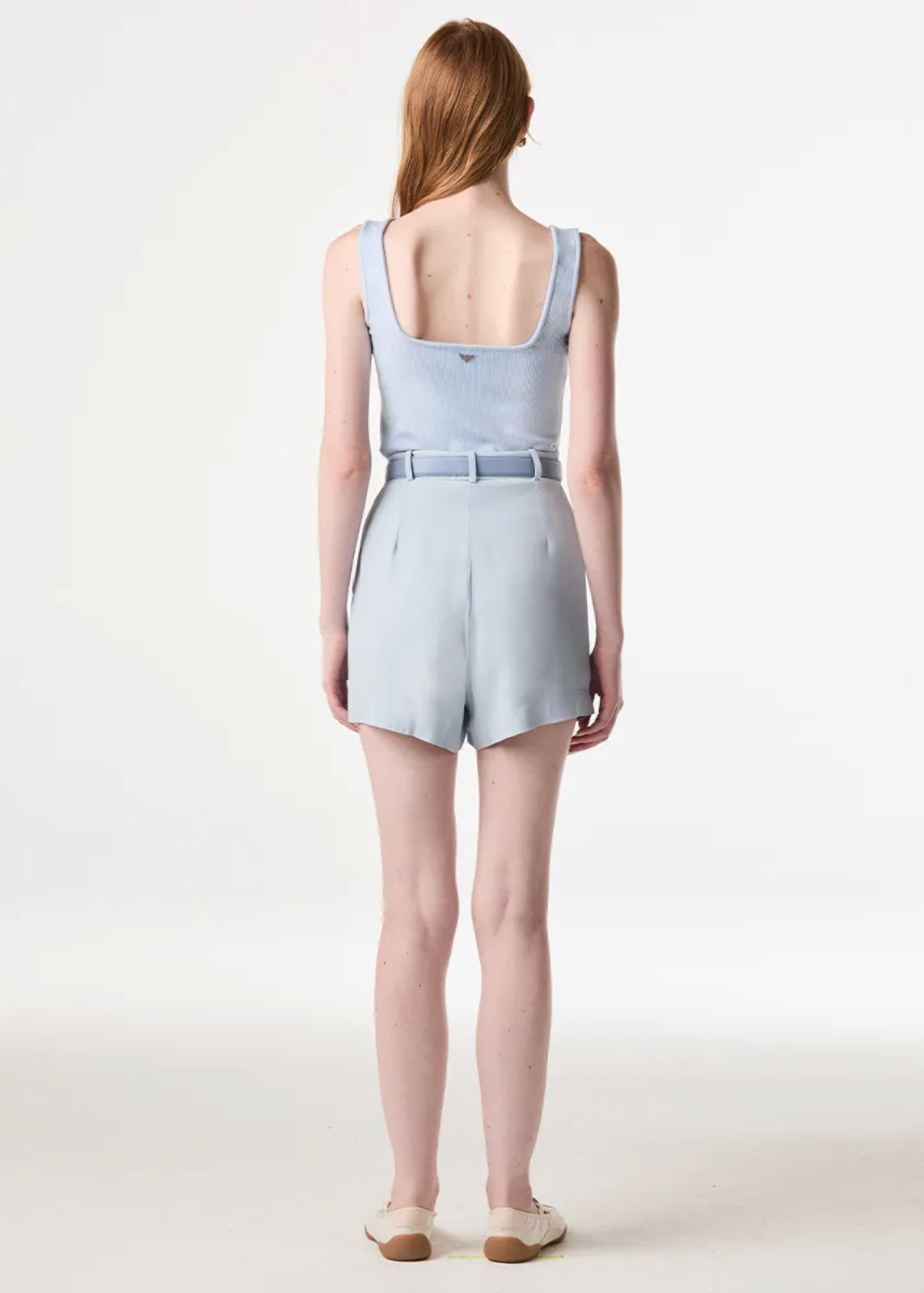 A model viewed from the back wearing a light blue sleeveless romper with a square neckline and a matching belt, set against a plain white studio background.