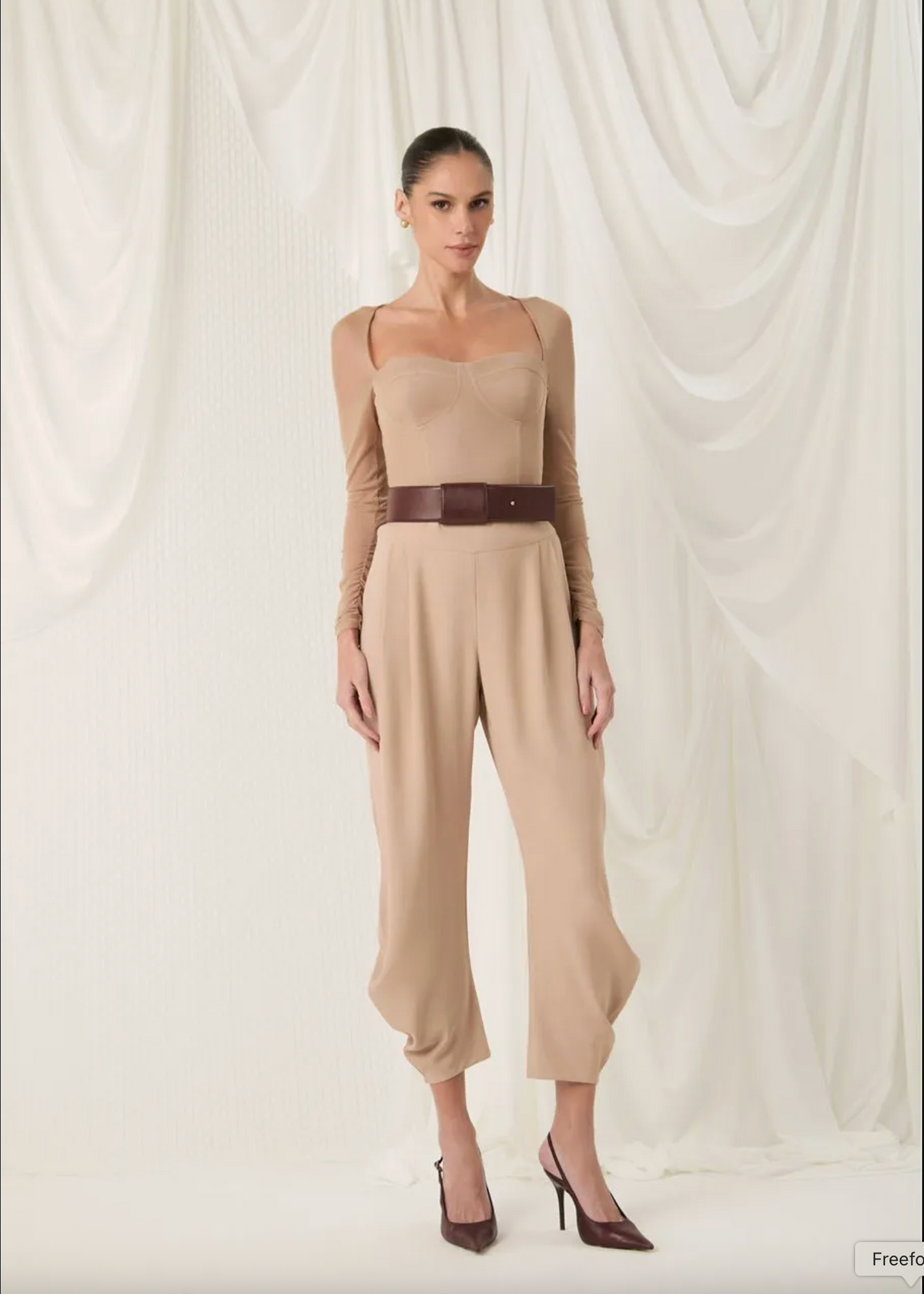 A model stands against a draped white fabric backdrop wearing a beige long-sleeve jumpsuit with a dark brown belt and matching dark brown heels.