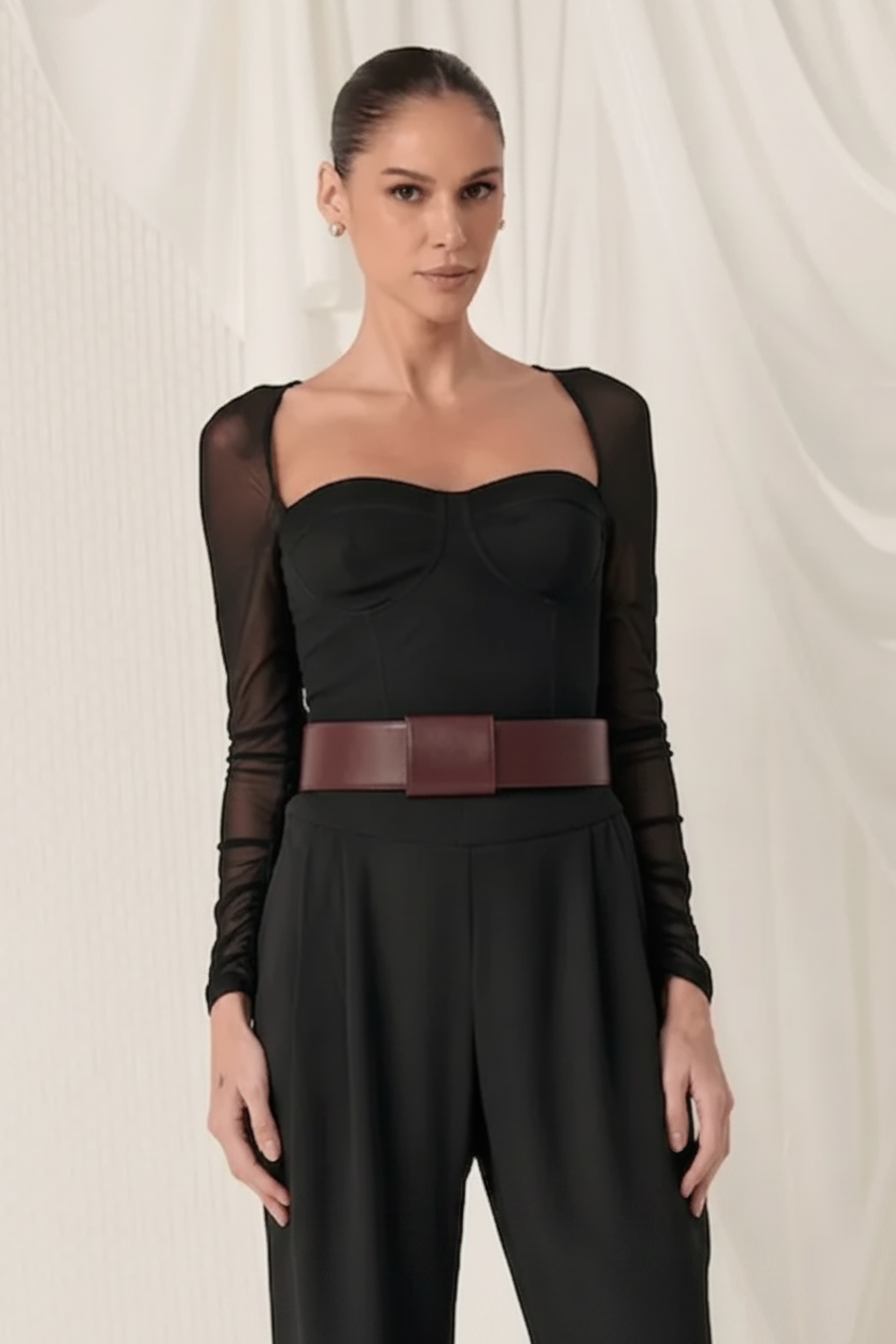 A woman modeling a black jumpsuit with sheer long sleeves, a sweetheart neckline, and a wide burgundy belt, posed against a draped white fabric background.