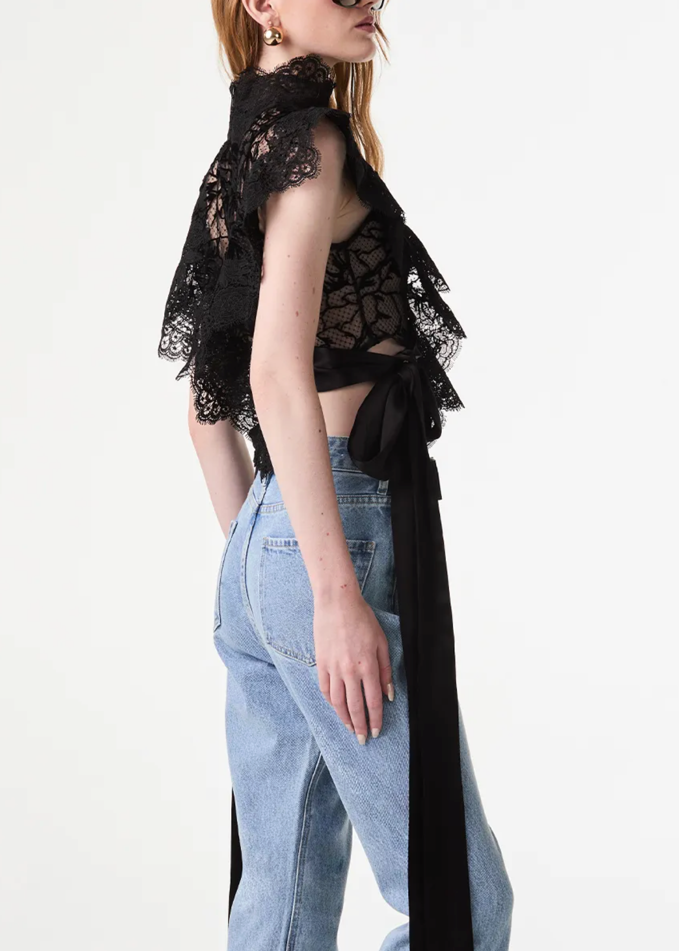 A model shown from the back wearing a black lace ruffled crop top with a long black ribbon tie and light-wash blue jeans, against a plain white studio background.